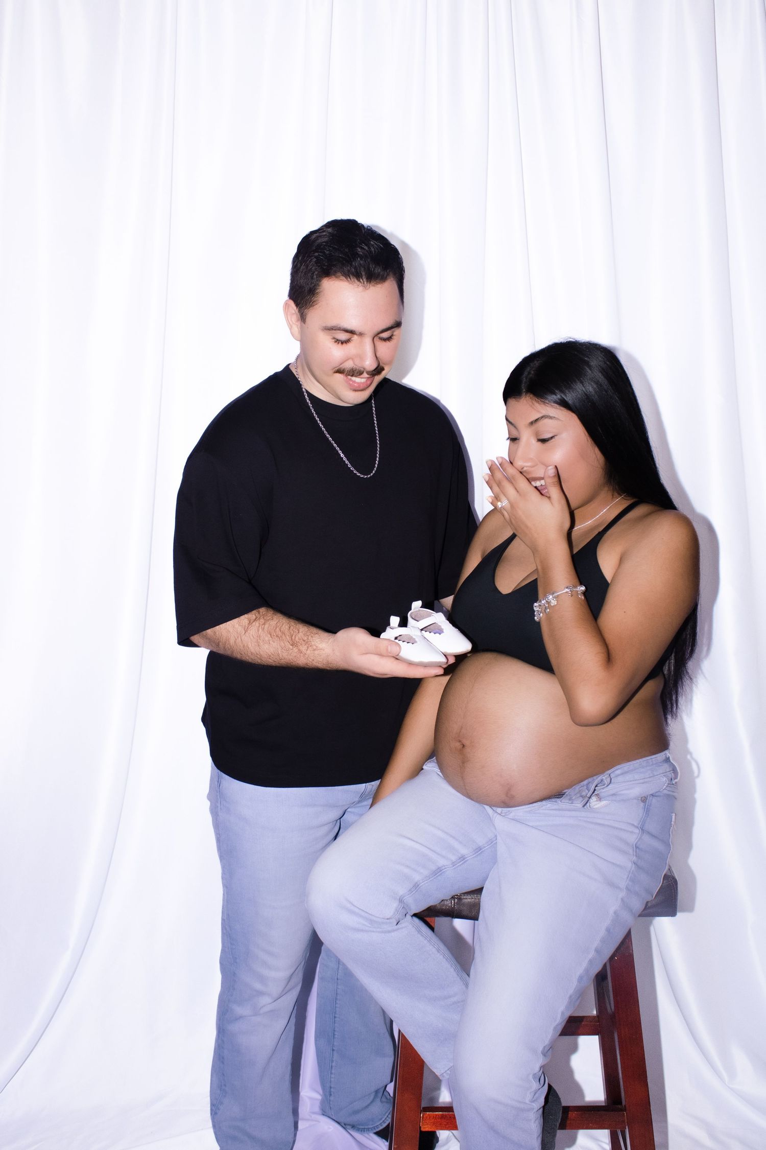 An expecting couple shares an emotional moment while holding tiny white baby shoes.