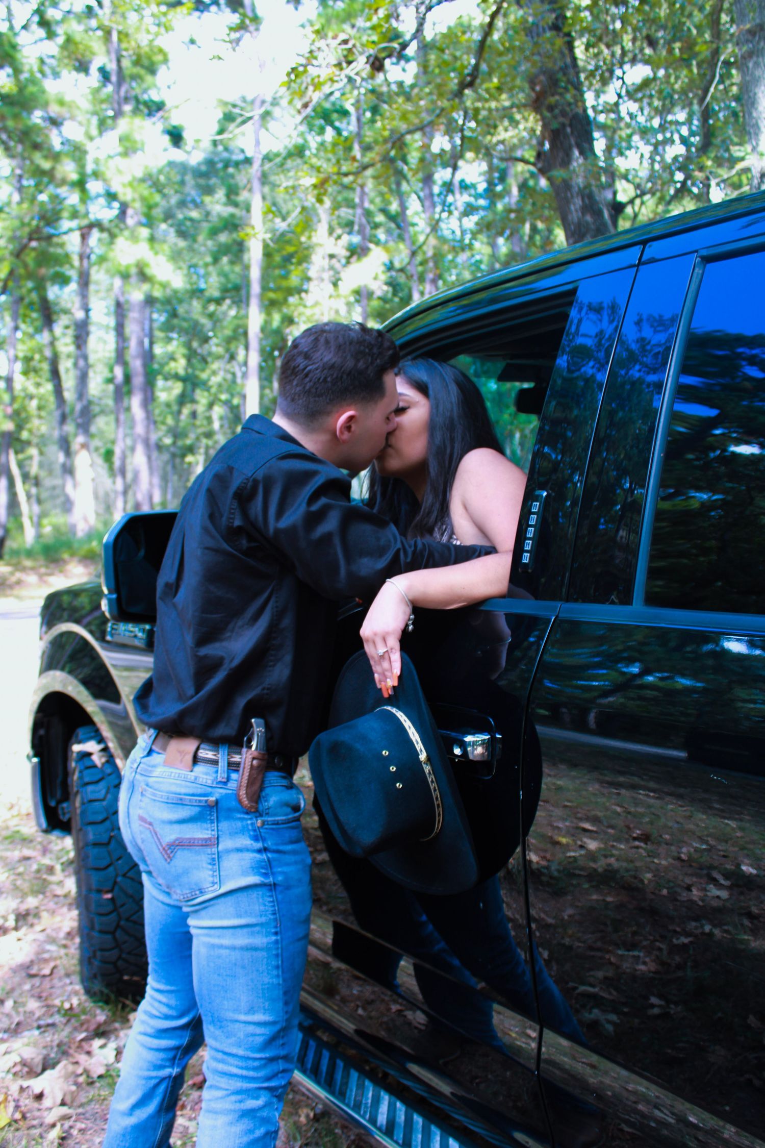 A couple leans against a black SUV parked in a wooded area.