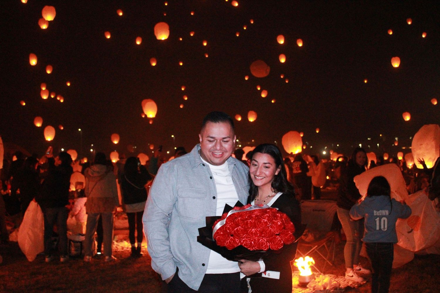 Couple celebrating with red roses under floating lanterns at nighttime festival.