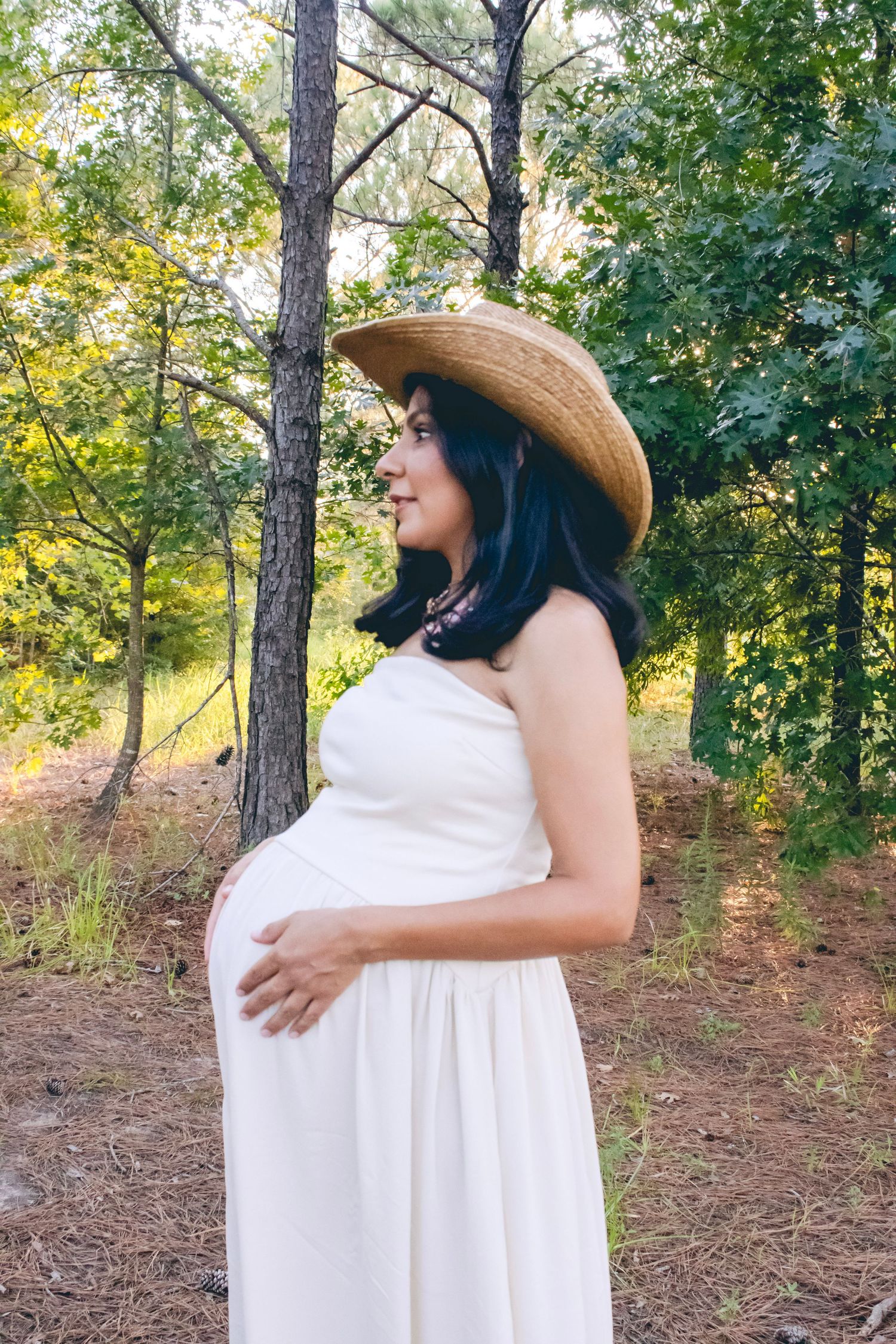 A maternity photo featuring a white dress and cowboy hat in a rustic outdoor setting.