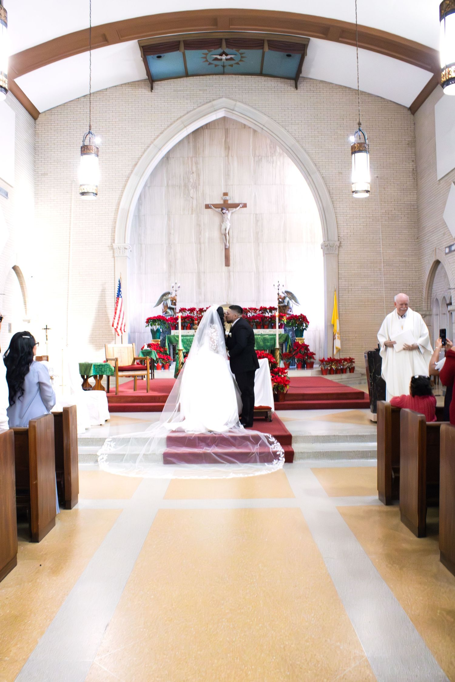 A wedding ceremony taking place in a church with pendant lights and wooden pews.