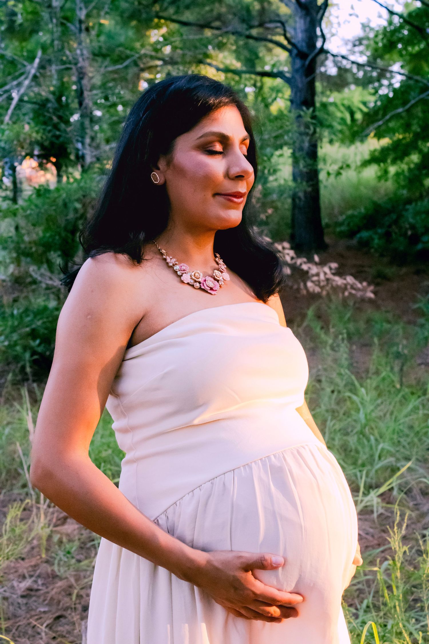An expectant mother in a cream-colored dress poses against a natural woodland backdrop.