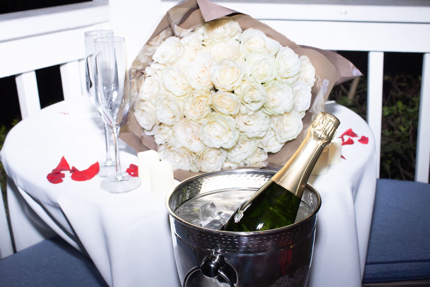 White roses and champagne in an ice bucket arranged on an outdoor table with red petals.
