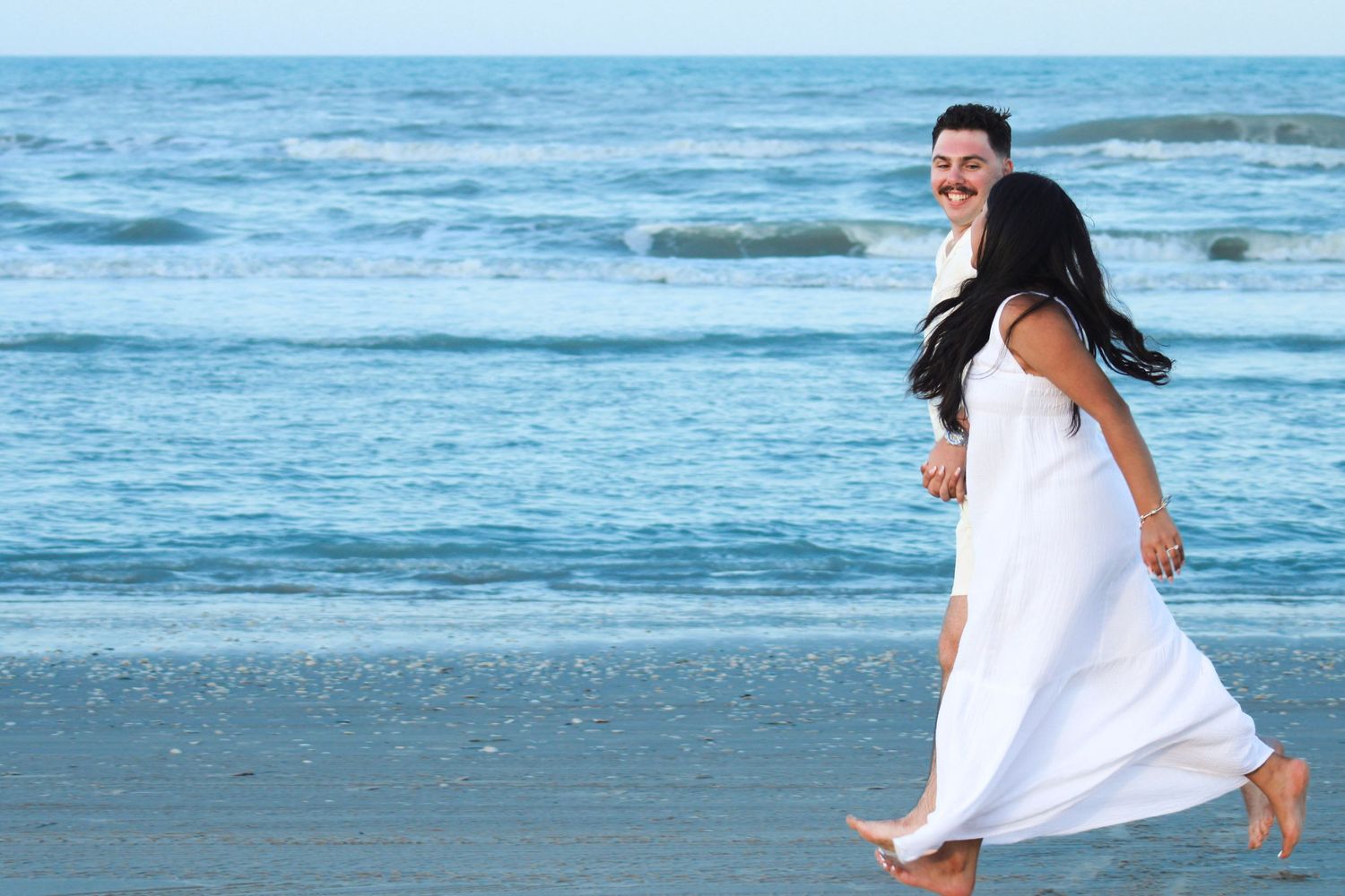 Couple in white outfits walking together on beach with ocean waves behind them.
