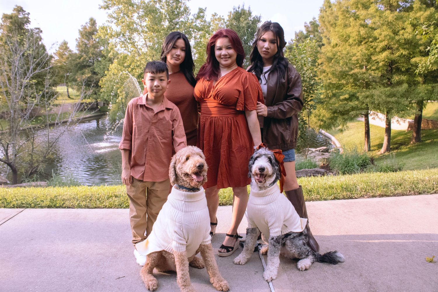 Two doodle dogs wearing cream sweaters pose with their owners in a park setting with autumn trees and a fountain.