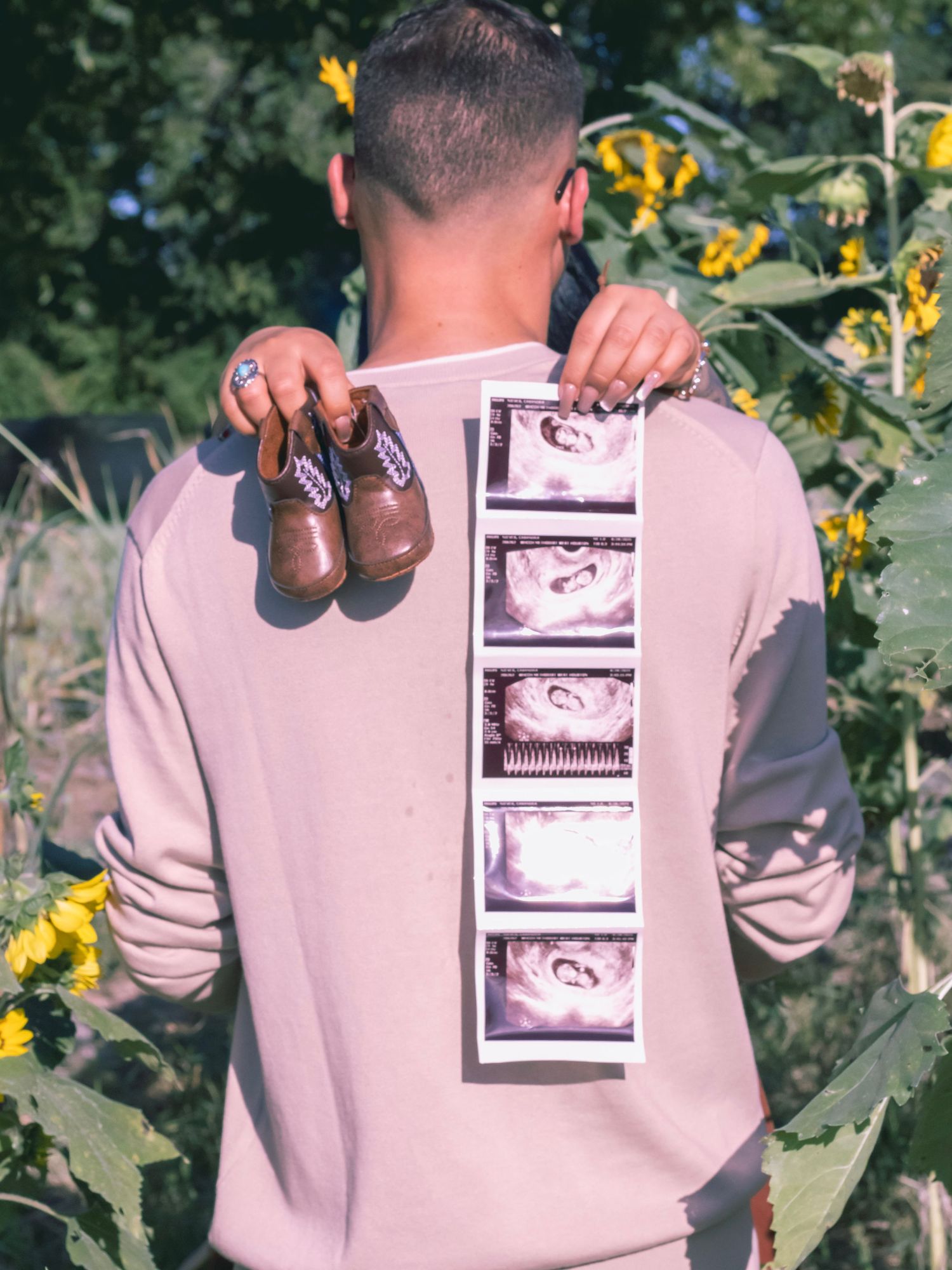 Baby shoes and ultrasound photos displayed on a person wearing a beige sweater among sunflowers.
