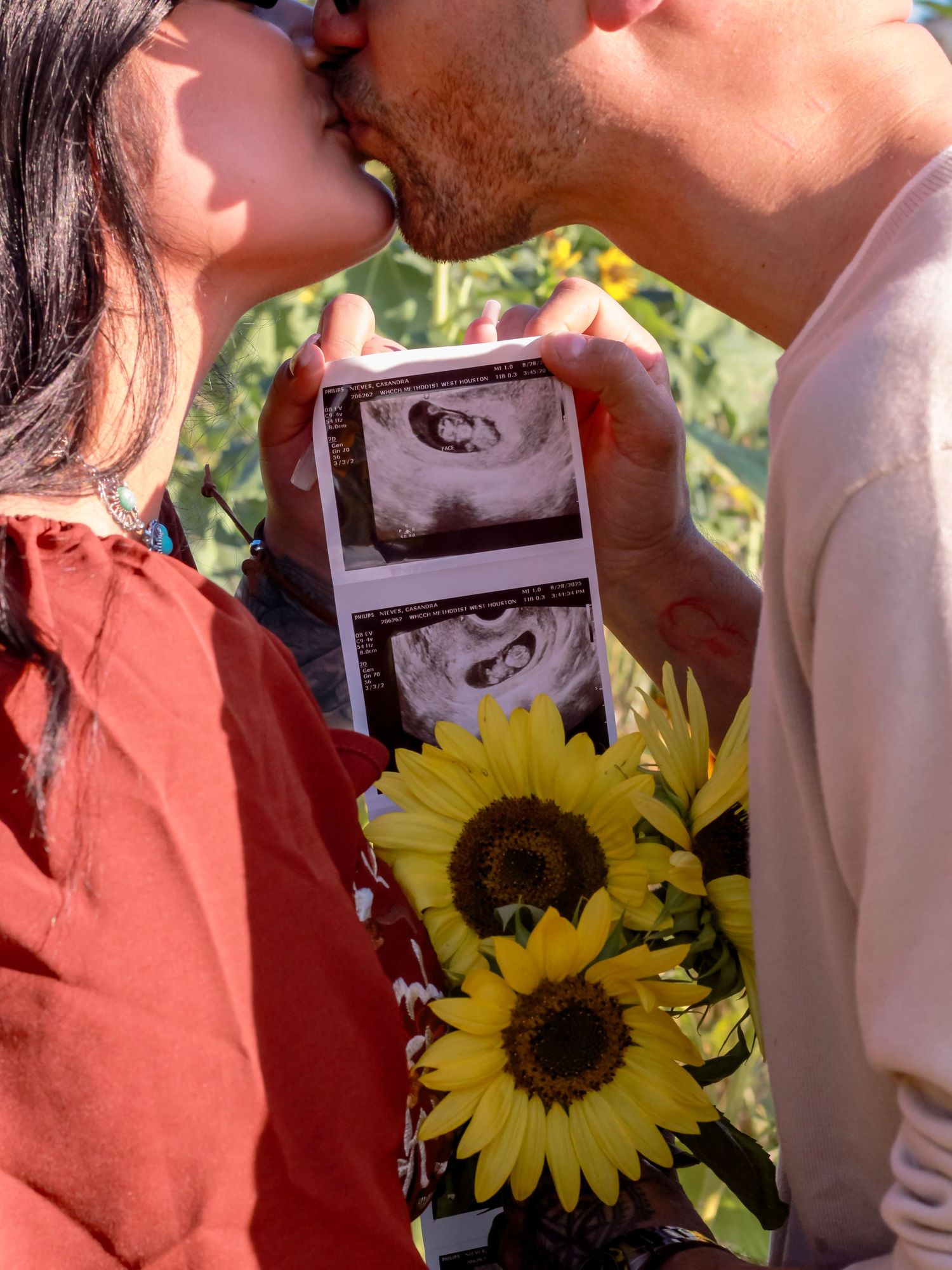 A couple shares a kiss while holding ultrasound photos and sunflowers in natural lighting.