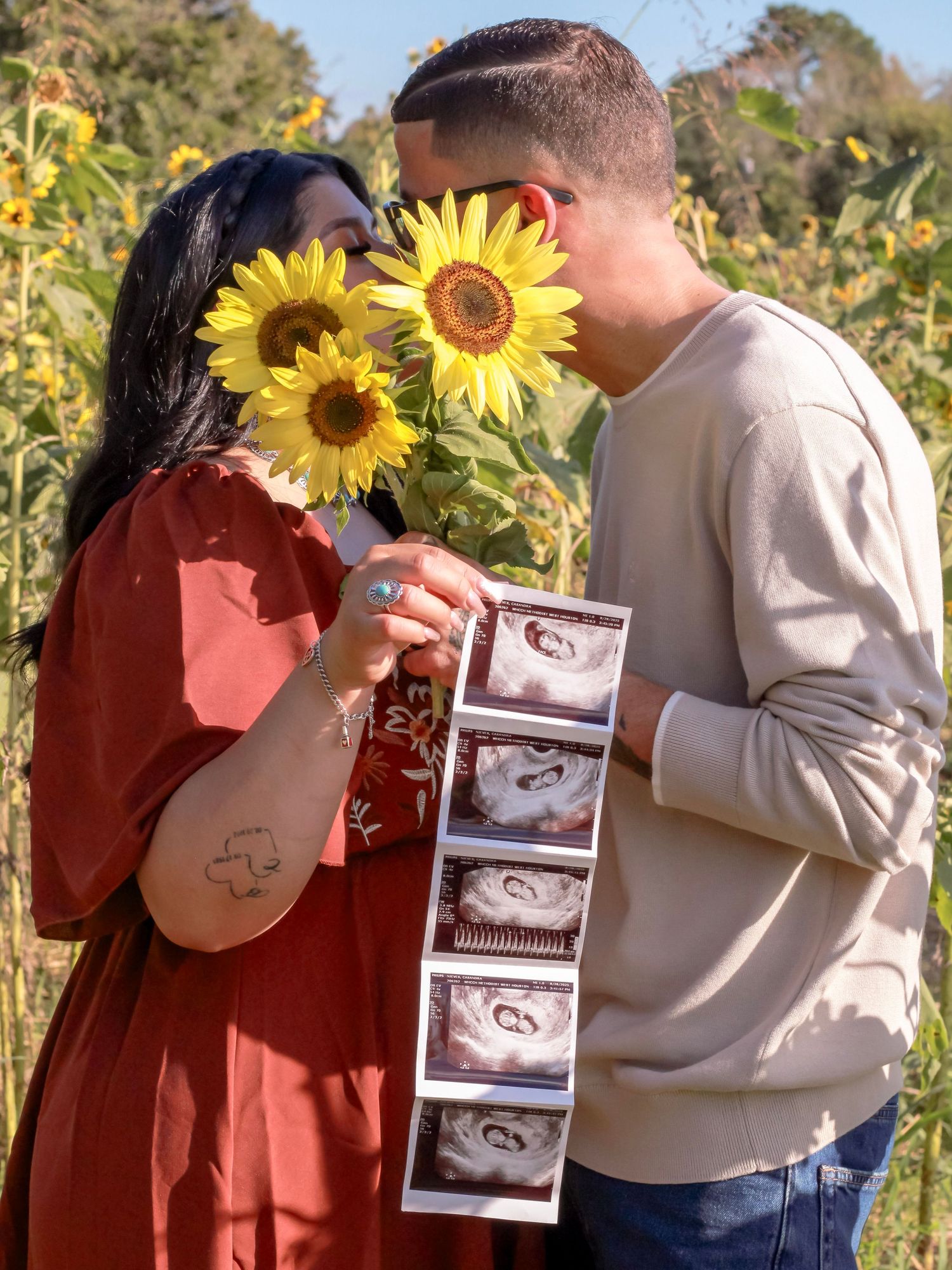 A couple poses with sunflowers and ultrasound photos in a golden sunflower field.