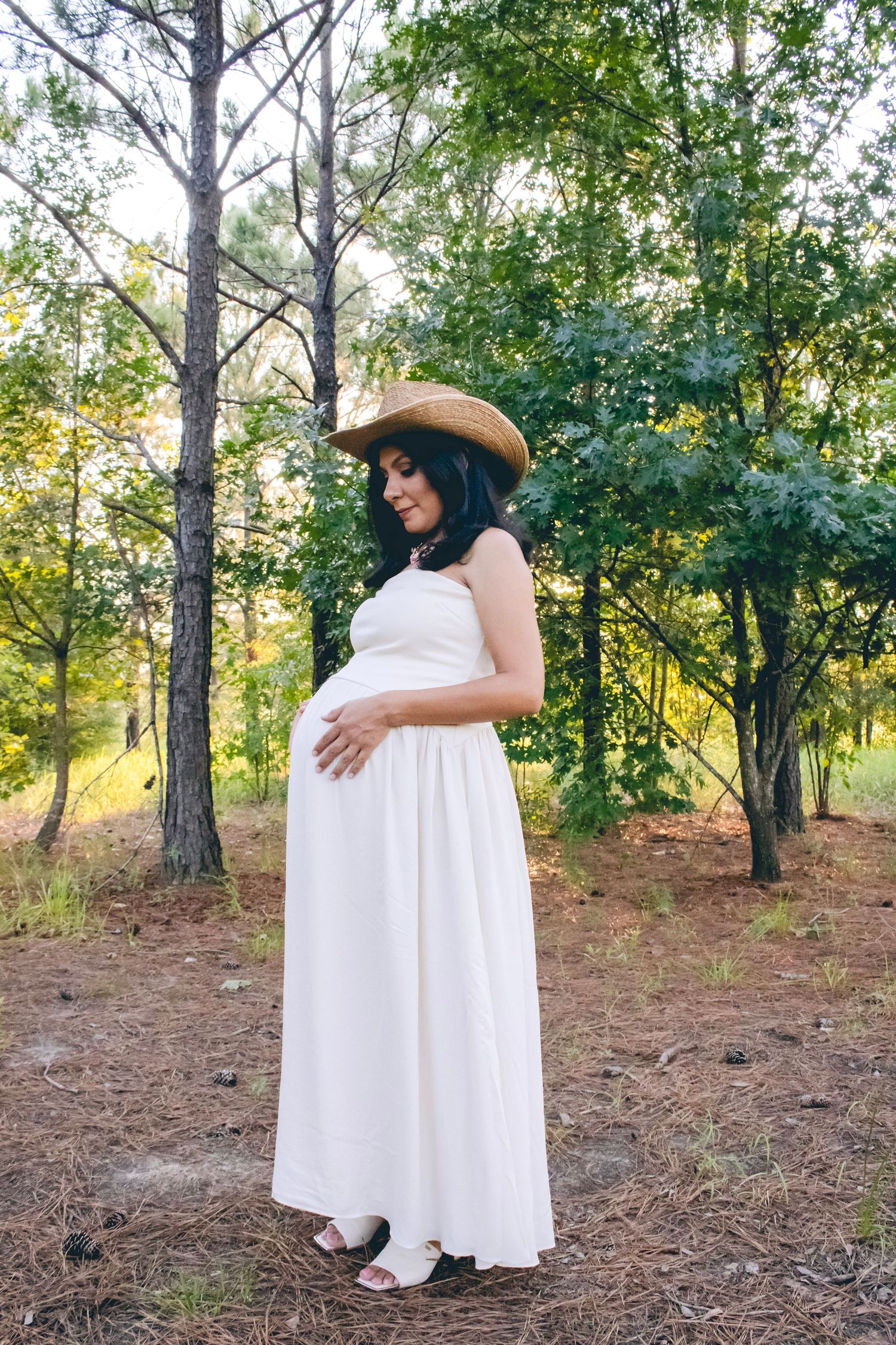 A pregnant woman in a white dress and sun hat stands among pine trees.