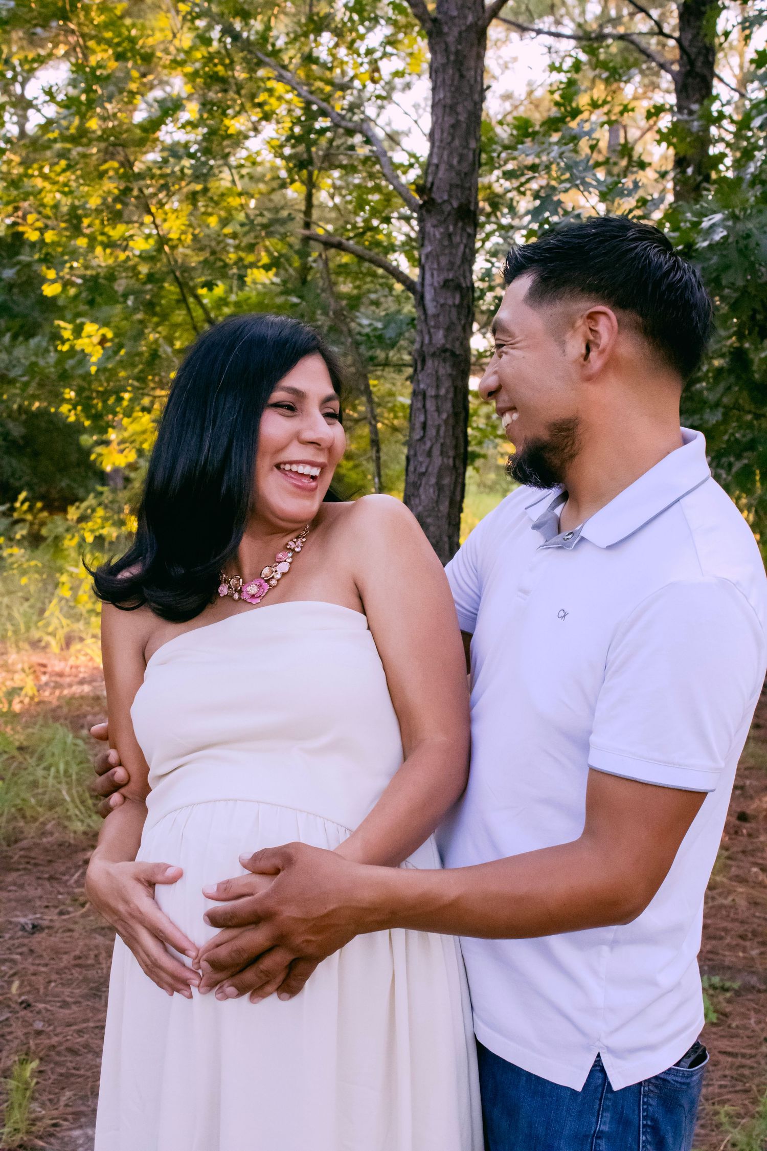 An expectant couple shares a joyful moment in a wooded setting during golden hour.