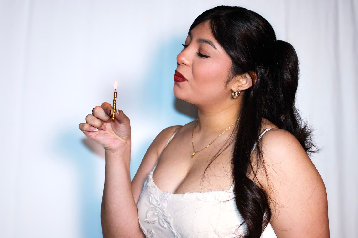 A person in a white dress poses playfully with a birthday cake against a light background.
