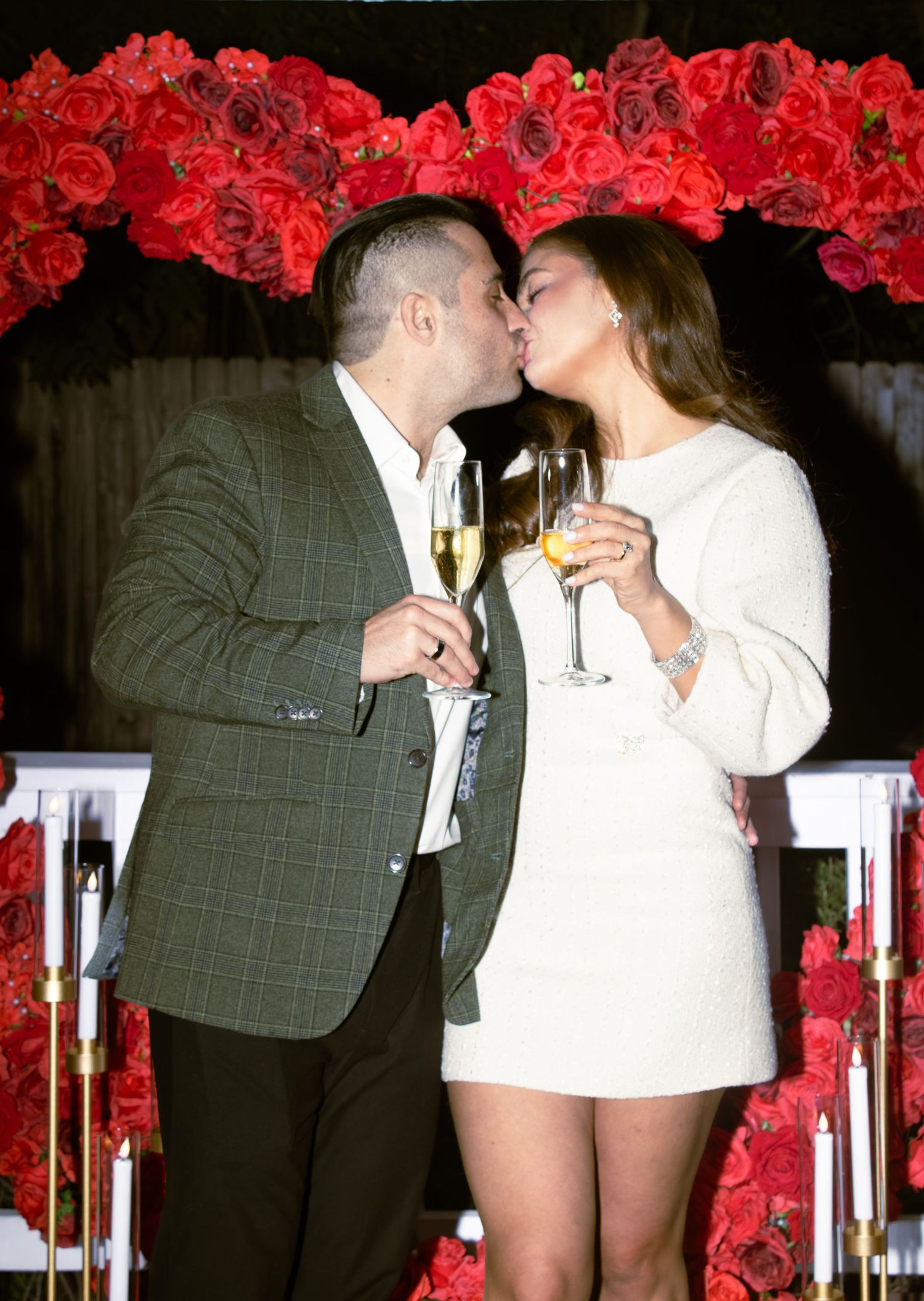 A couple shares a kiss under a red rose arch while holding champagne glasses.