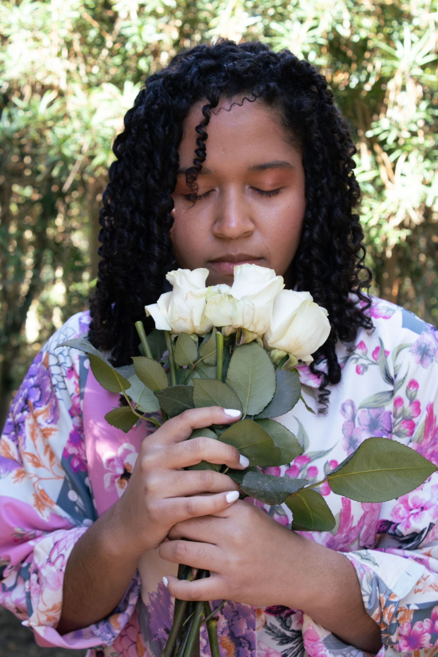 Someone smelling white roses while wearing a pink floral dress in a natural outdoor setting.