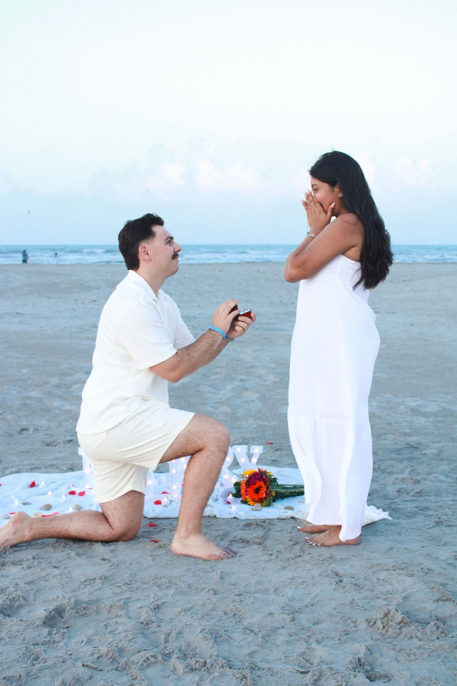 Marriage proposal scene on beach with person kneeling in white attire.