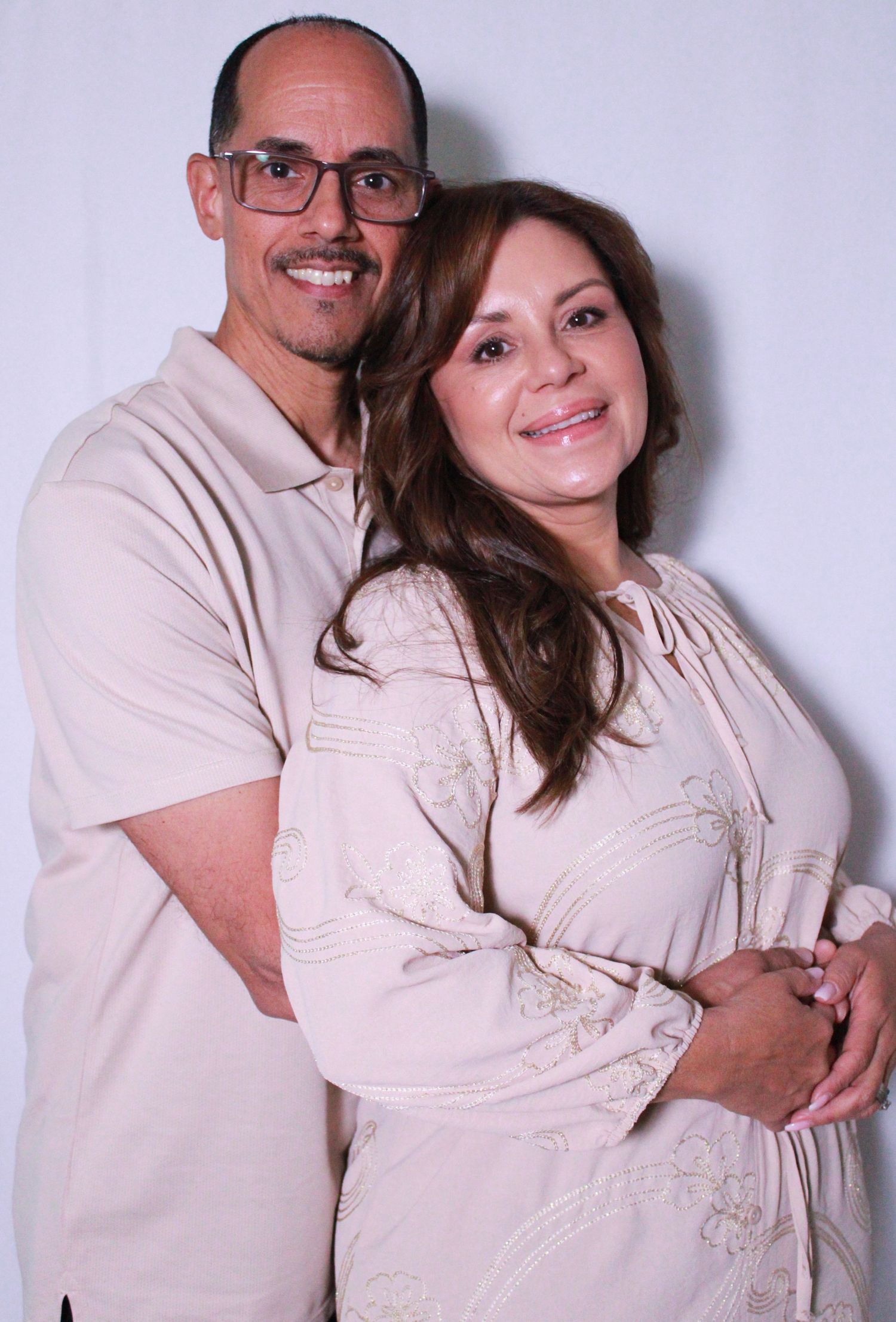 A couple poses together against a light gray studio backdrop wearing coordinating beige outfits.