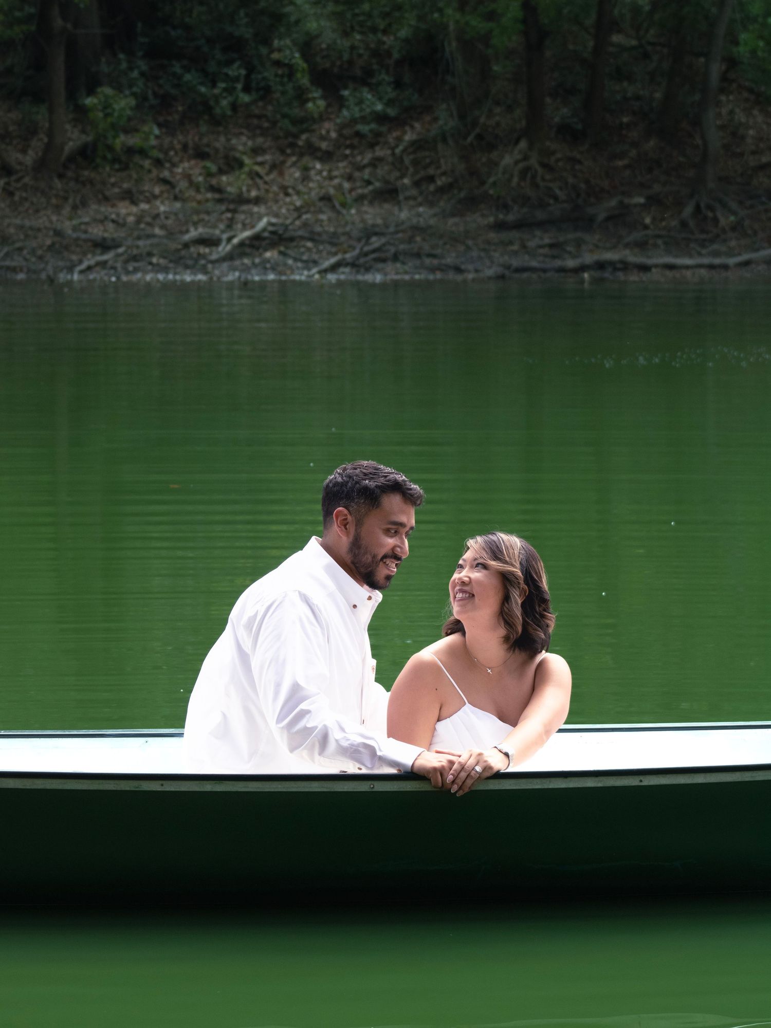 A couple shares a joyful moment while sitting in a white boat on a green lake.