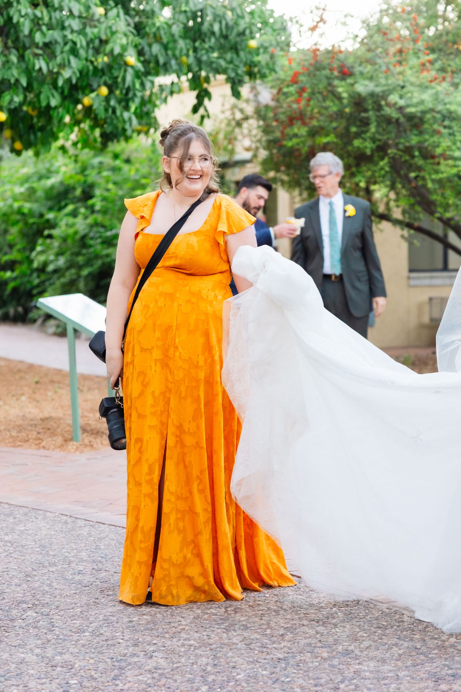 Bridesmaids in bright orange dresses walk alongside a bride in a flowing white gown on a garden path.