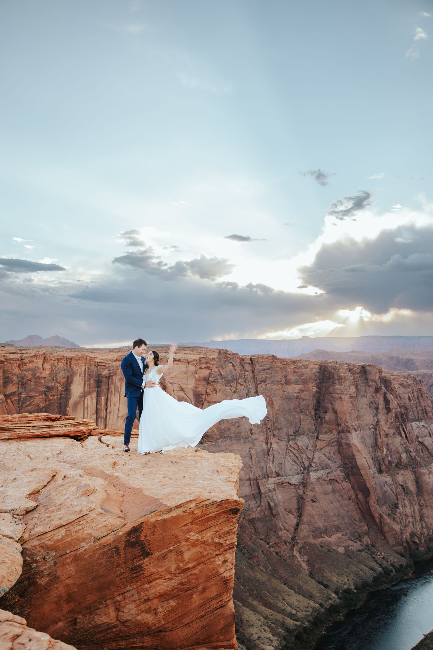 Couple in wedding attire poses dramatically on red rock cliffs overlooking scenic desert canyon at sunset.