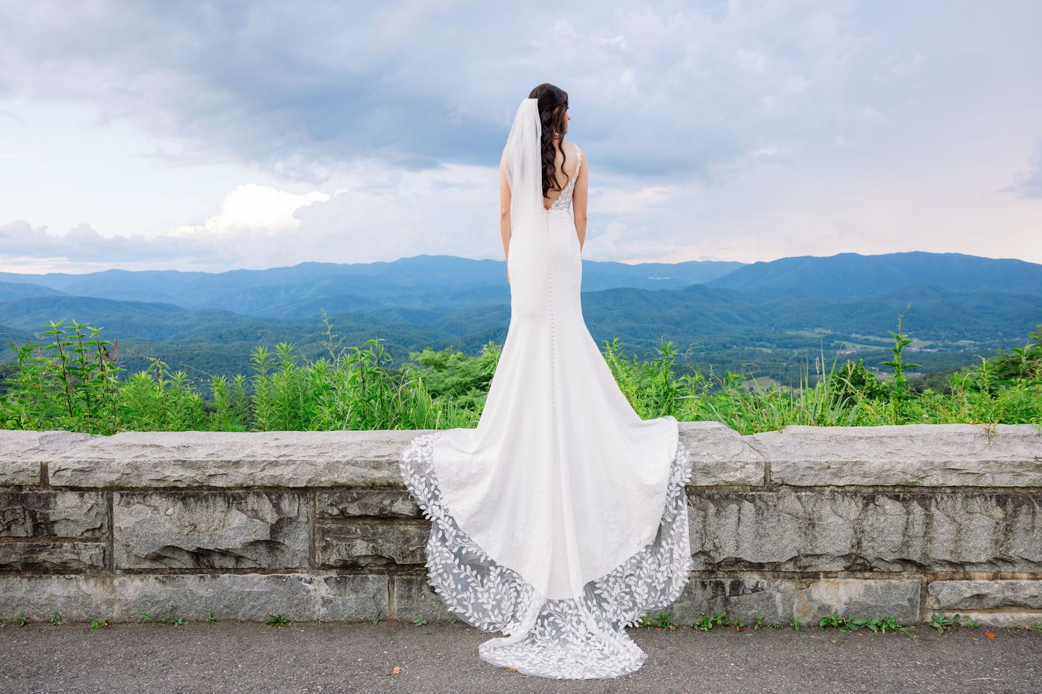 A wedding dress silhouette stands against a stone wall with mountains and green landscape in the background.