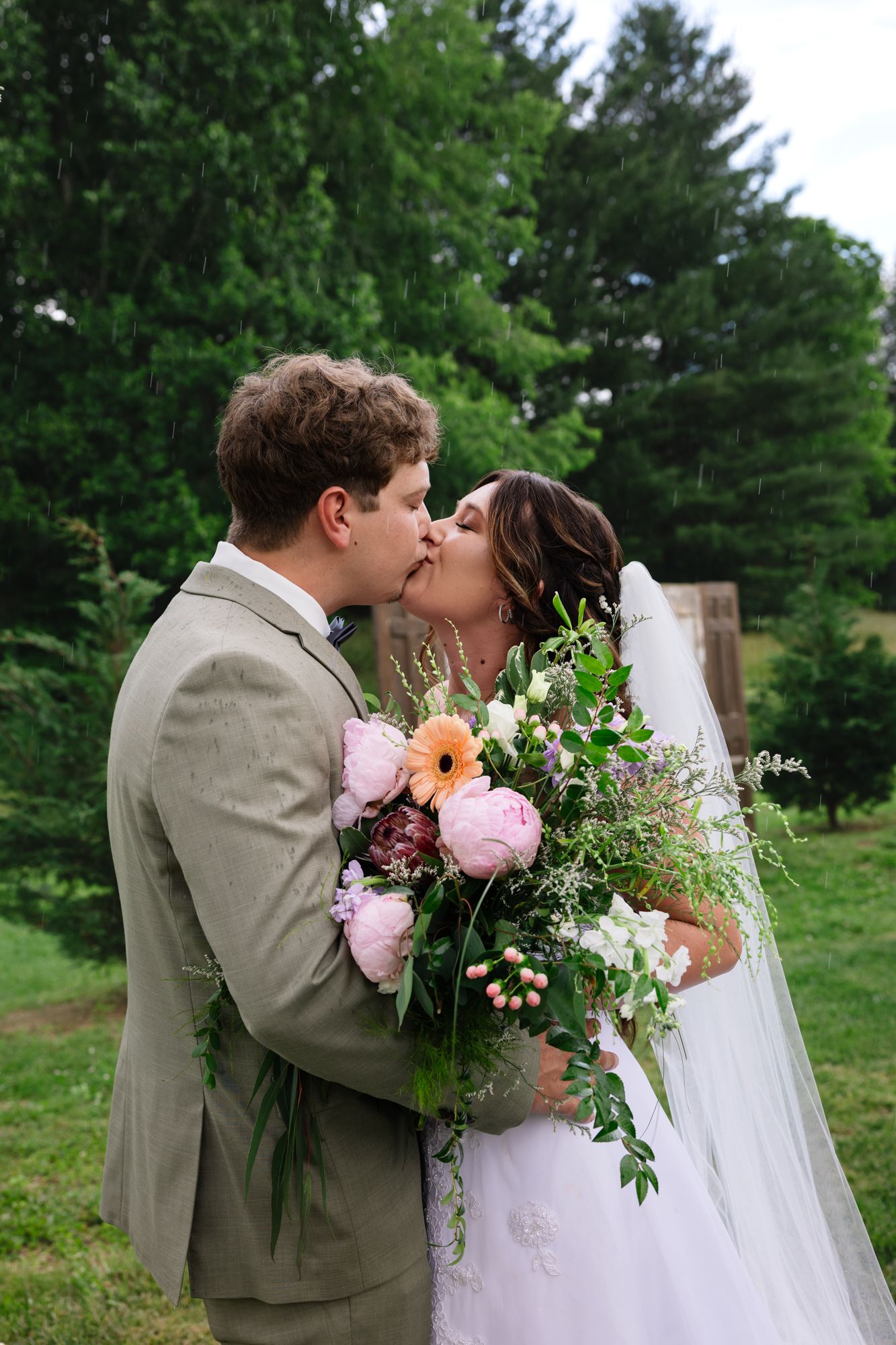 A couple in wedding attire share a romantic kiss outdoors surrounded by beautiful green trees and holding floral bouquets.