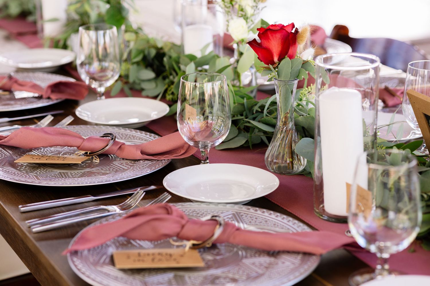 Elegant table setting with pink napkins, red roses in bud vases, white candles and greenery garland on wooden table.