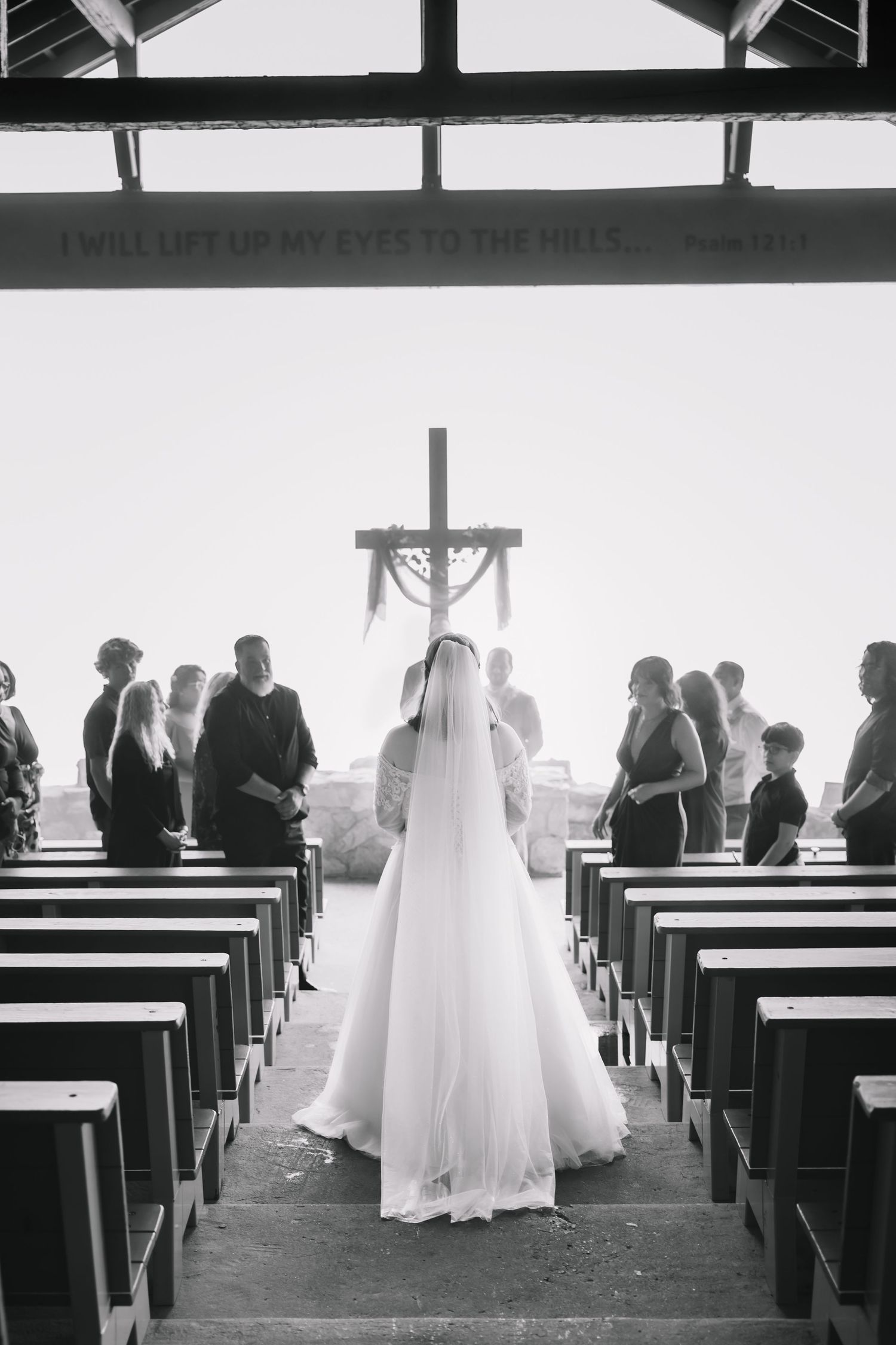 Black and white photo of a bride in white gown standing at church altar with guests seated in wooden pews.