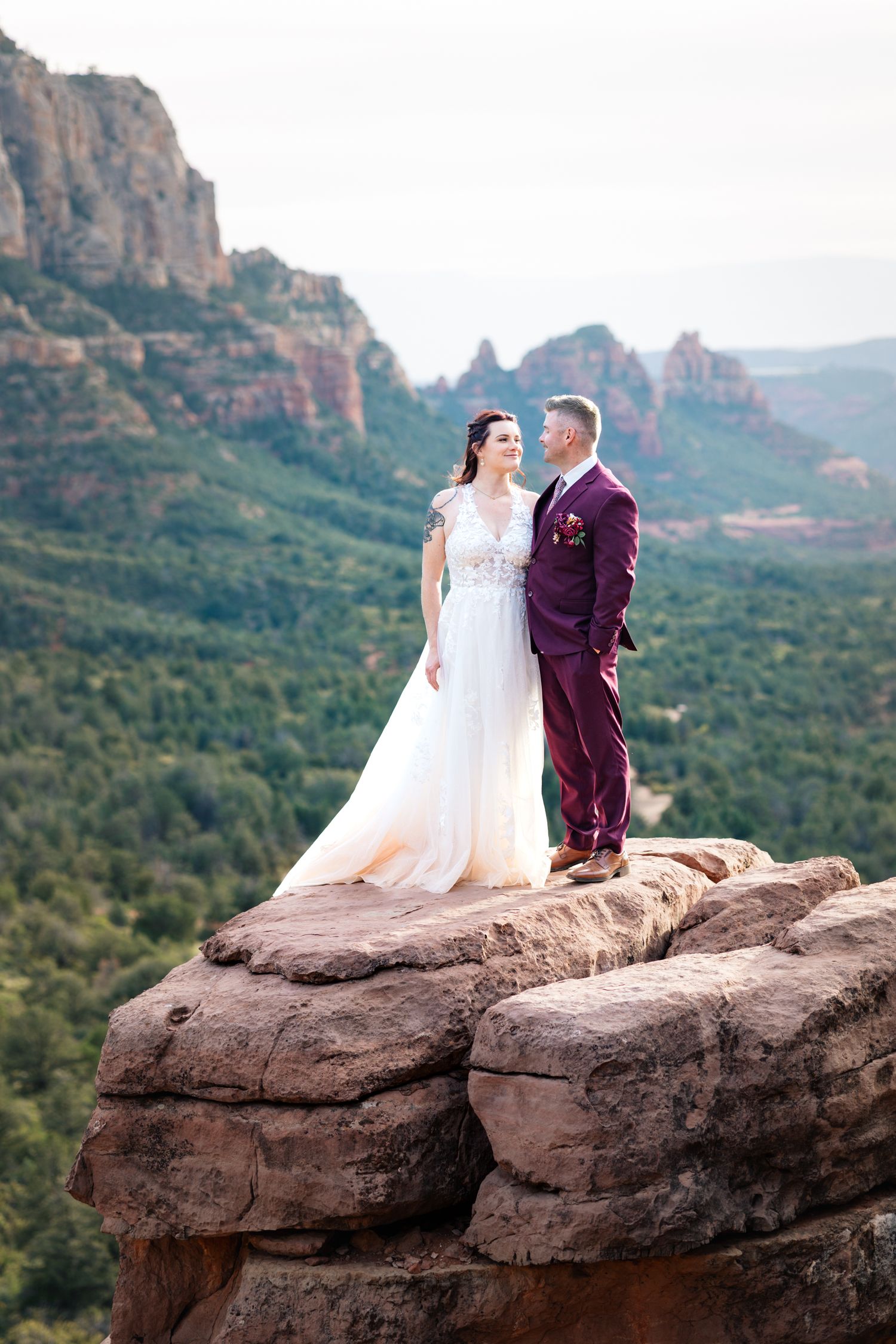 A couple in wedding attire poses on a red rock formation overlooking the scenic mountains of Sedona, Arizona.