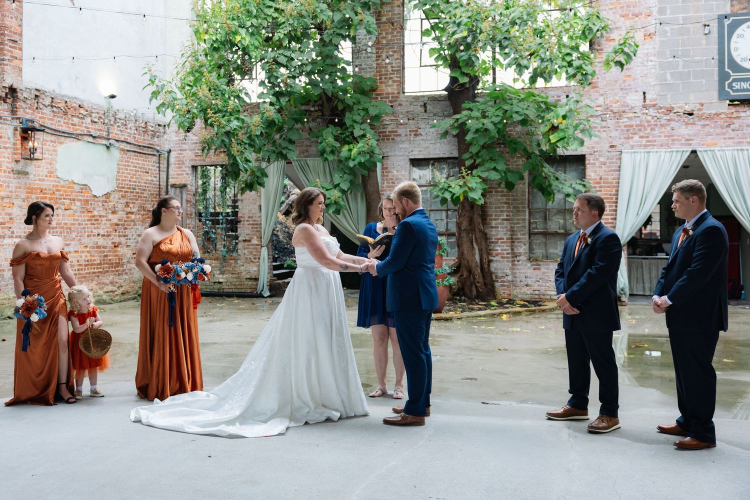 A wedding party stands together in a rustic brick courtyard during an outdoor ceremony.