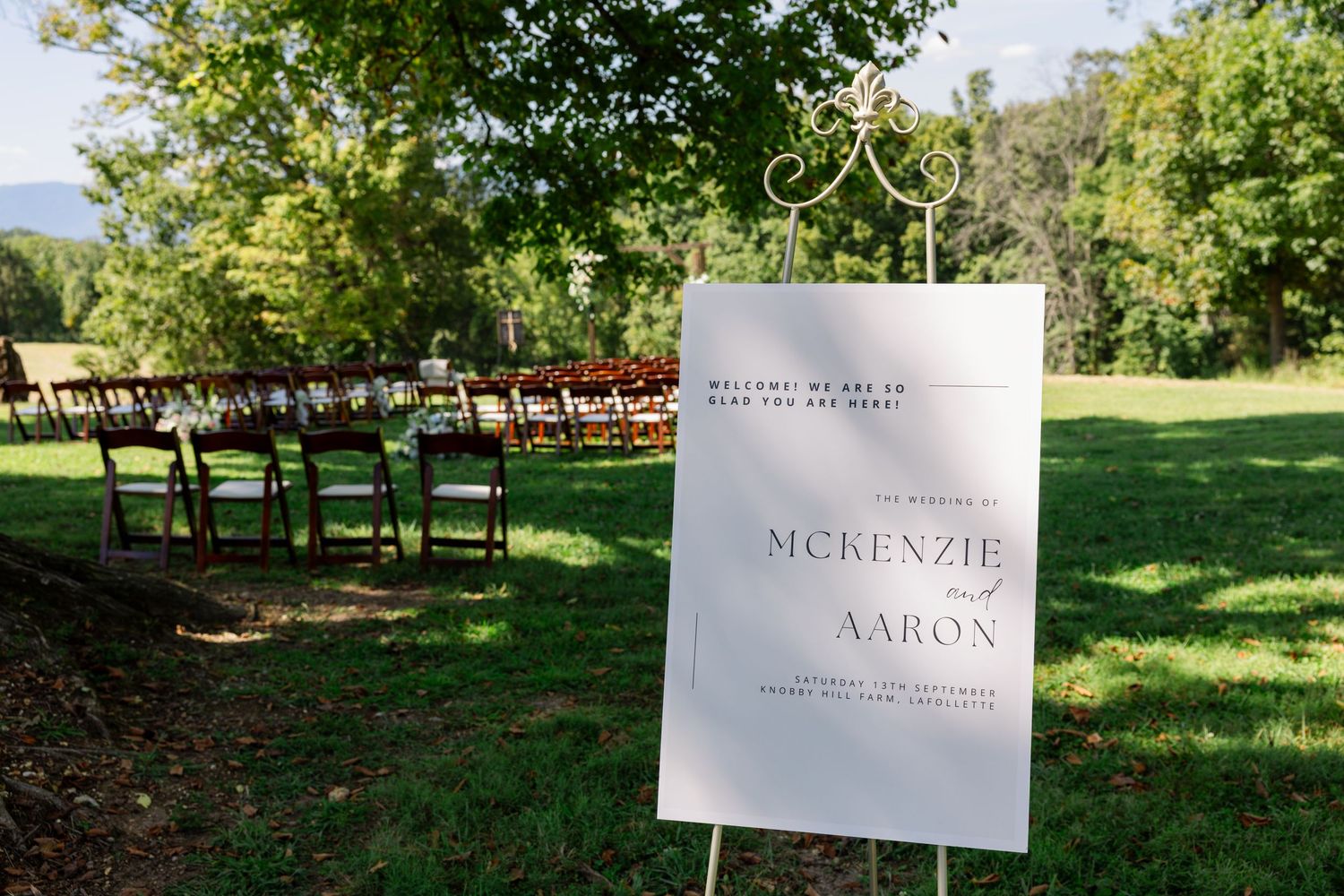 A scenic outdoor wedding ceremony setup with white chairs arranged in rows on a lush green lawn surrounded by trees.