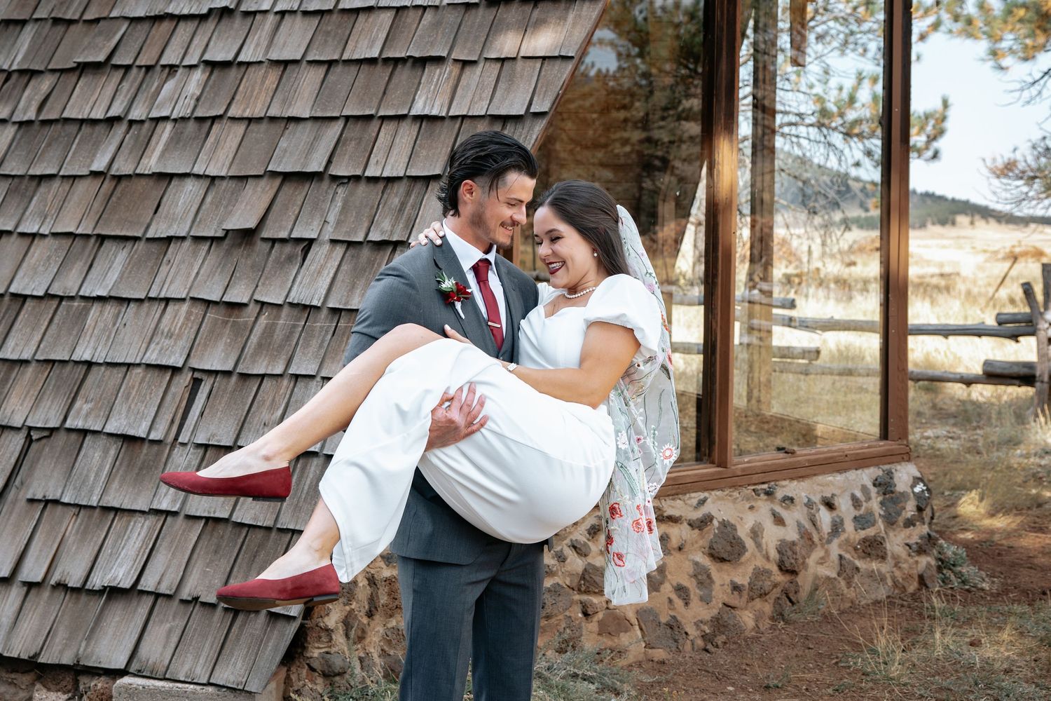 A romantic couple embraces outside a rustic church with cedar shingles as one carries the other.