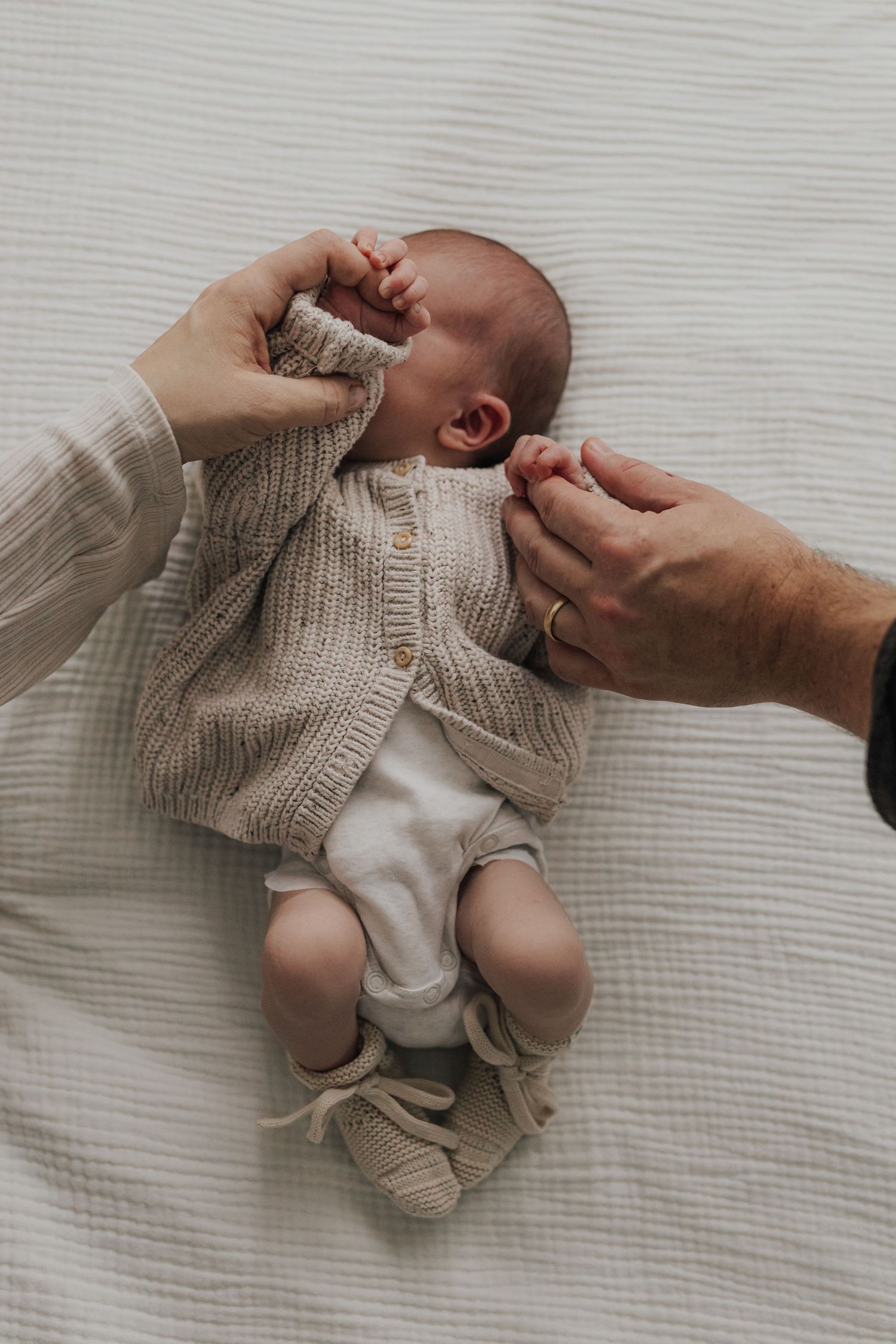 A sequence of hands gently dressing a small baby in a knit sweater on white bedding.