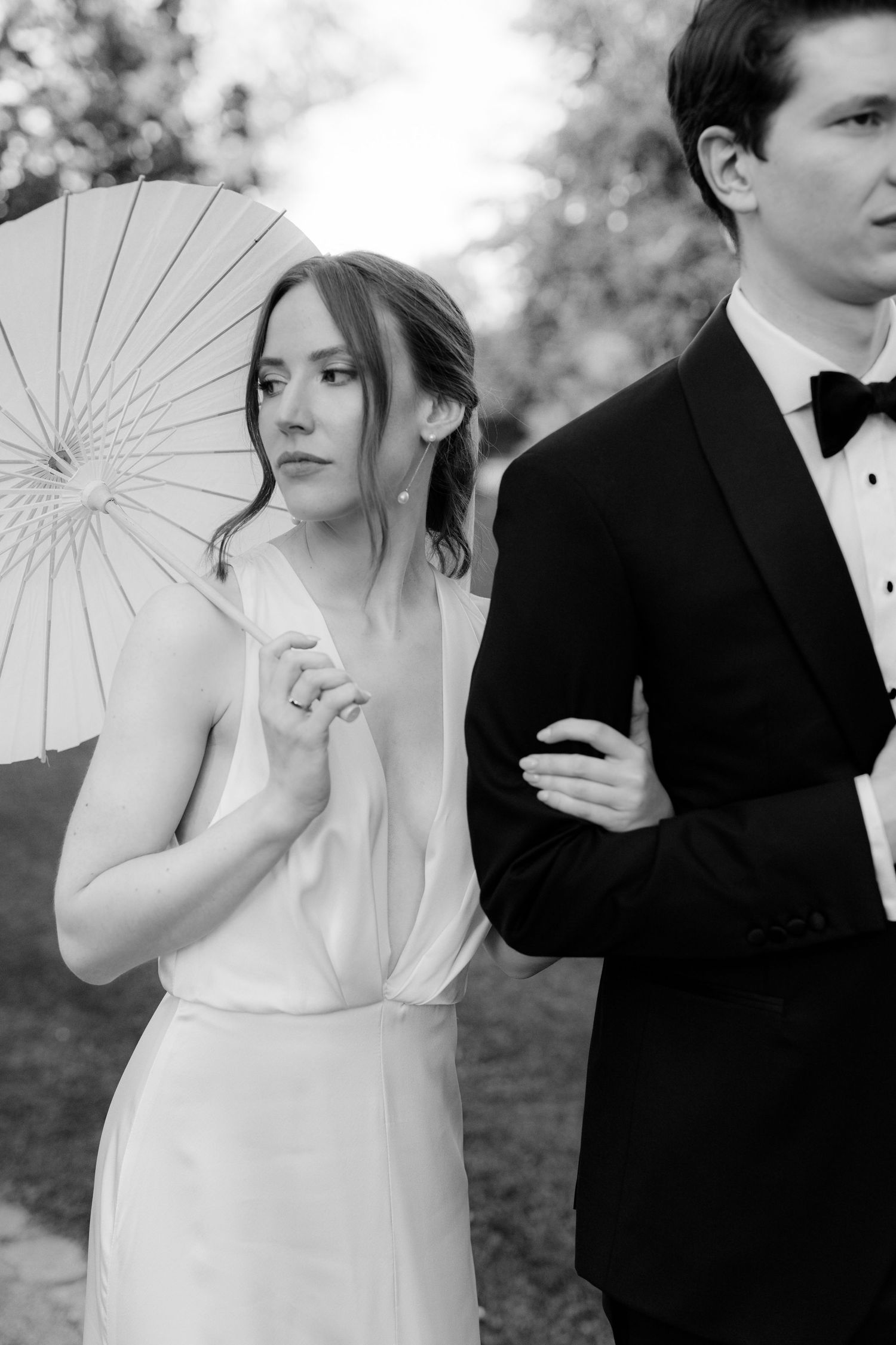 Black and white photo of a bride holding a parasol in a white gown, standing beside a groom in a tuxedo outdoors.