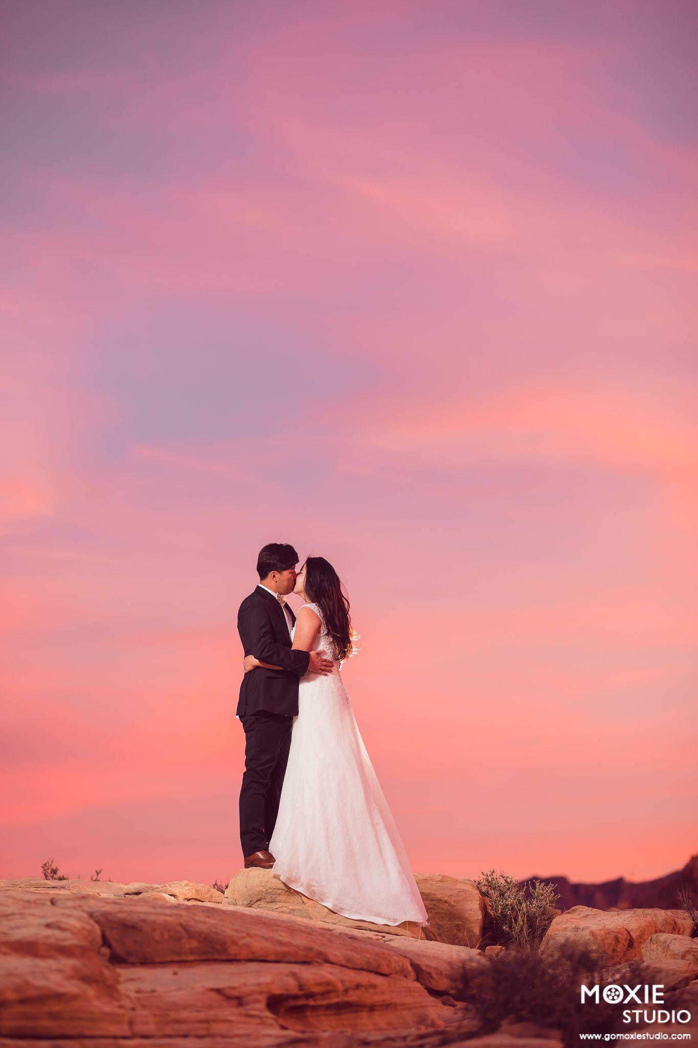 Wedding couple embracing on red rock formations during a dramatic pink and purple desert sunset.