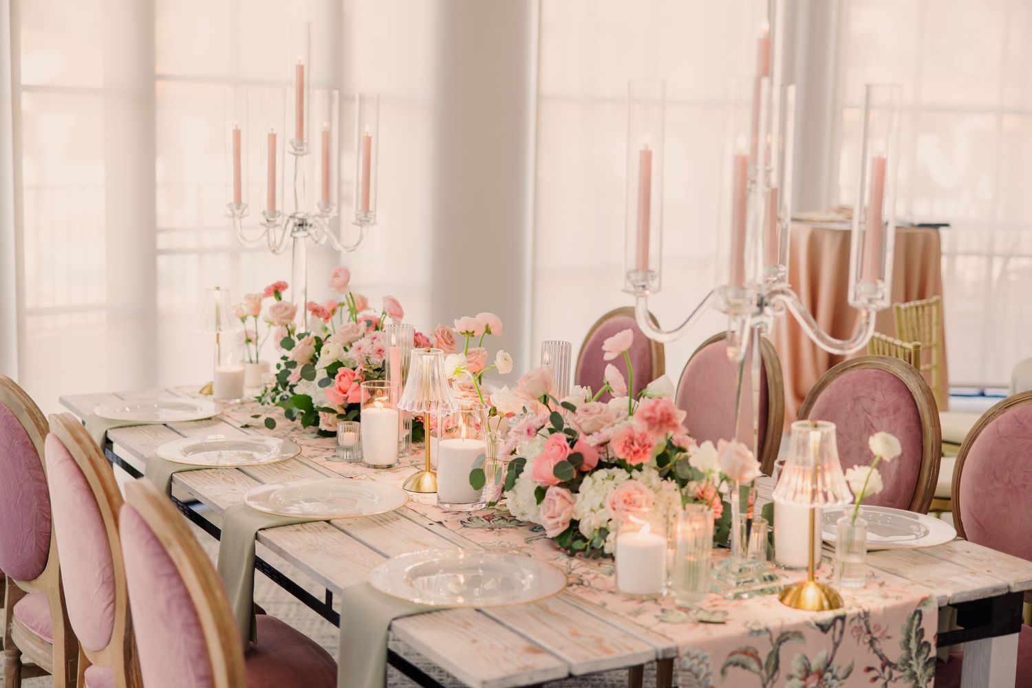 Elegant wedding reception table settings with blush pink linens, floral centerpieces and glowing candles in a bright venue.