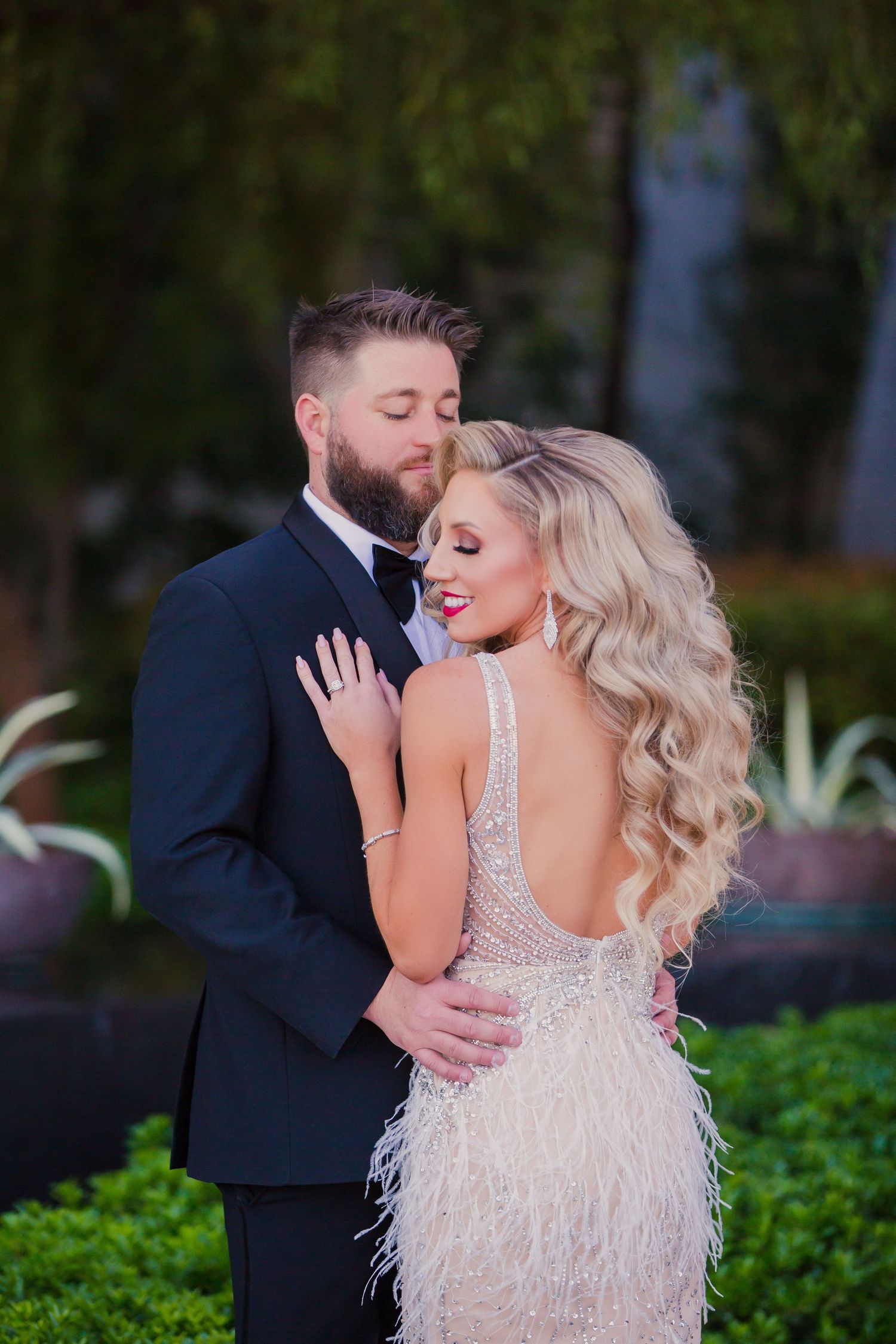 Couple embraces in garden setting with bride wearing sparkly backless dress with feathers and groom in navy suit. Red Rock Casino