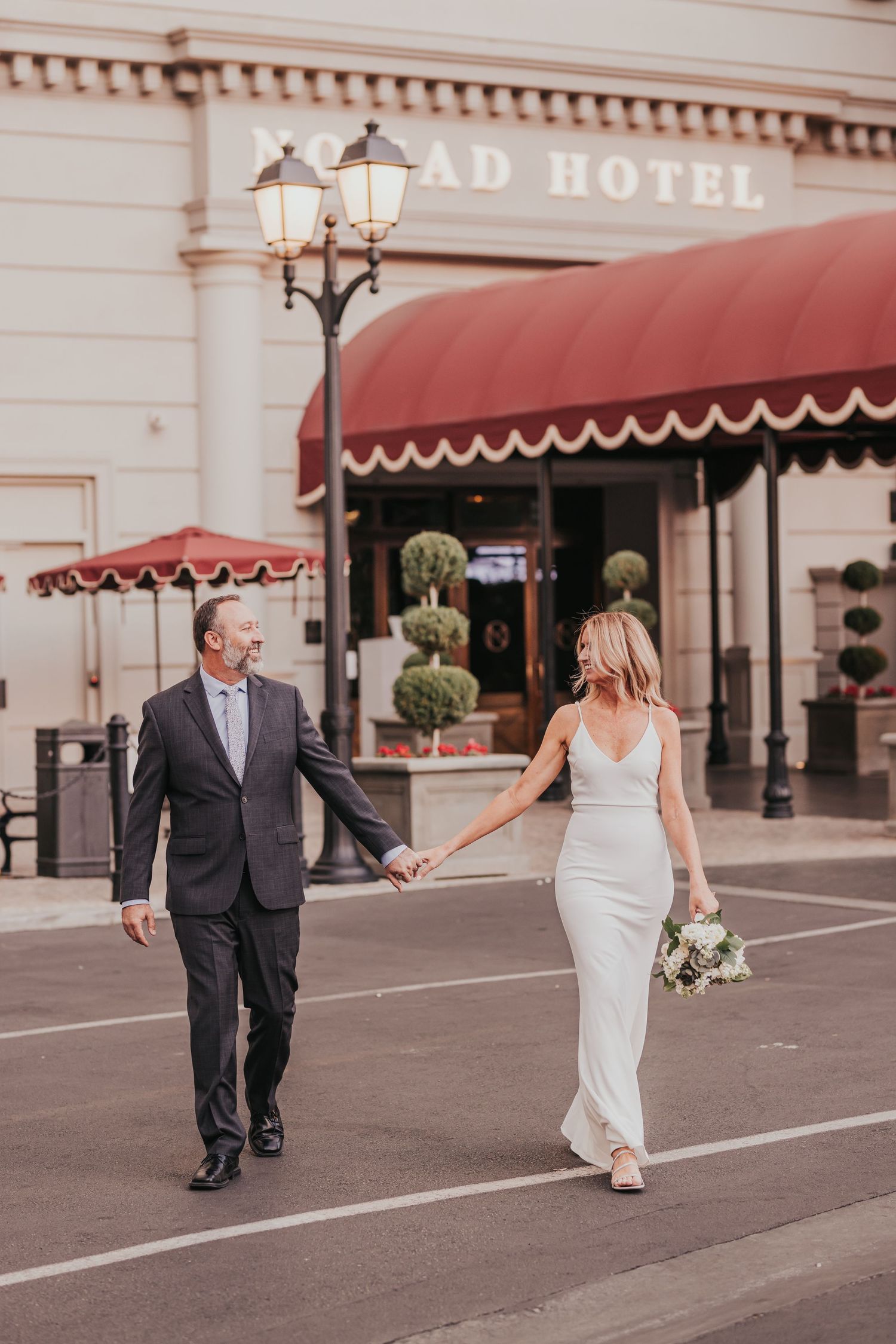 Newlywed couple walks hand in hand along a historic hotel exterior with red awnings in a romantic urban setting.
