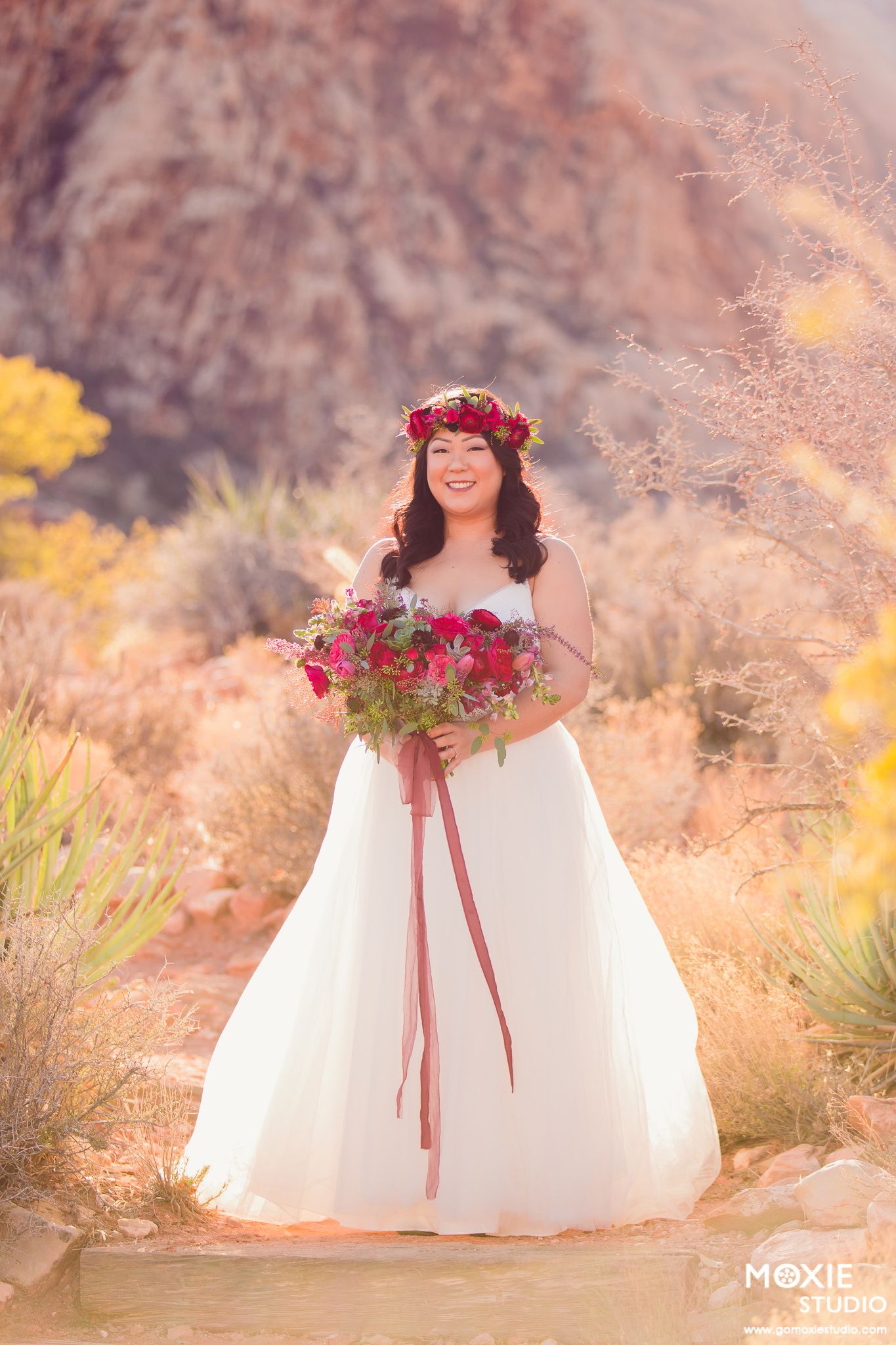 A person in a white wedding dress with red floral accents poses in a desert landscape at sunset.