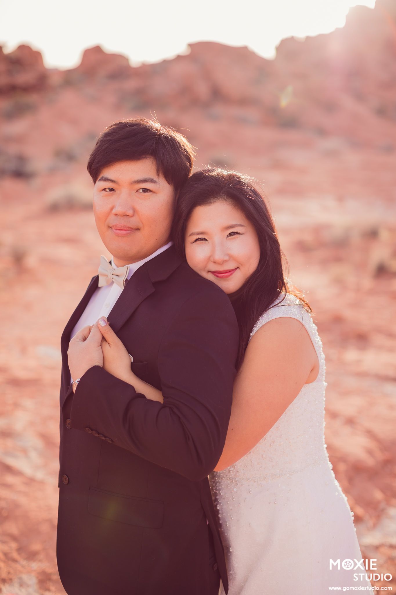A couple in wedding attire embrace while posing in front of red rock formations in a desert landscape.
