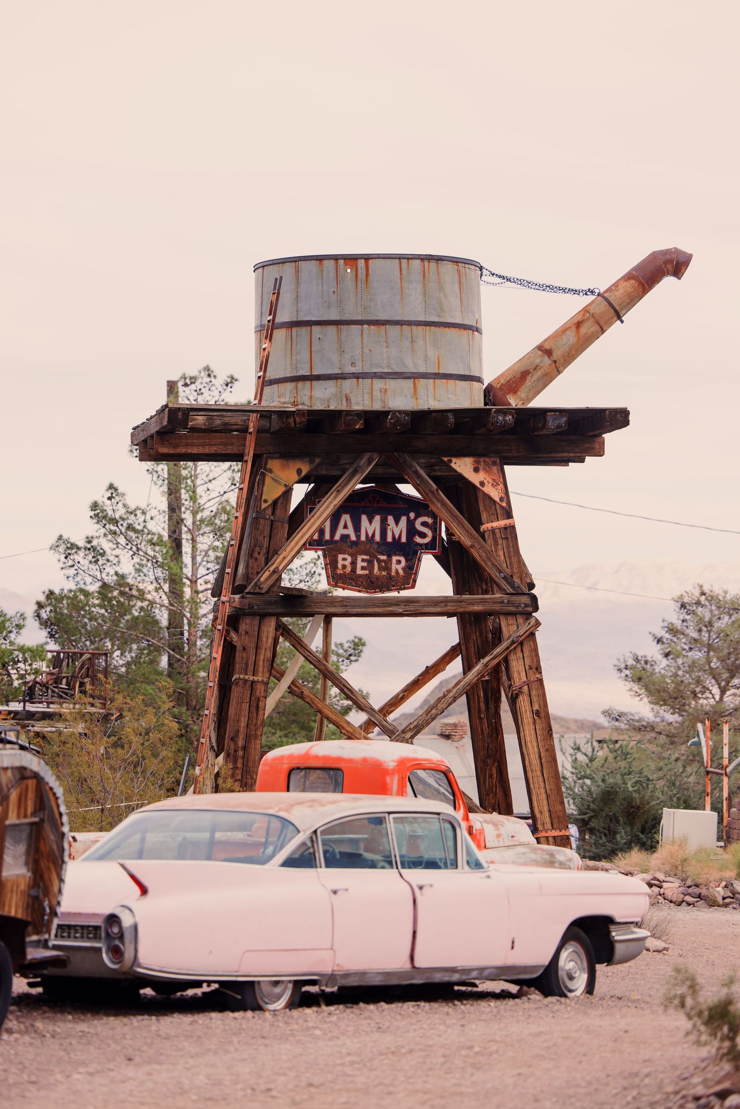 Vintage cars parked beneath rustic wooden water towers with Hamm's Beer signage in a desert setting.