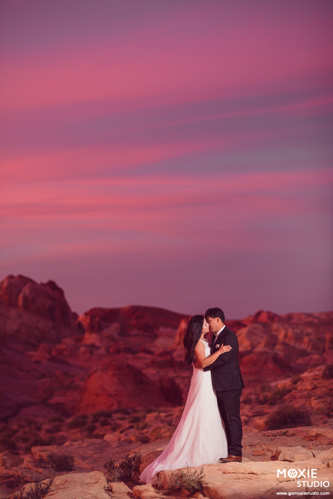 Wedding couple shares an intimate moment against dramatic purple desert sunset and rocky terrain.