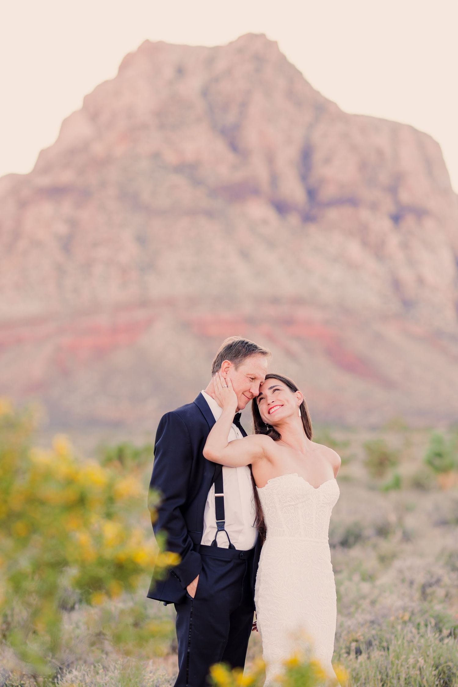 A romantic couple embraces in front of majestic red rock mountains in a desert landscape during sunset.