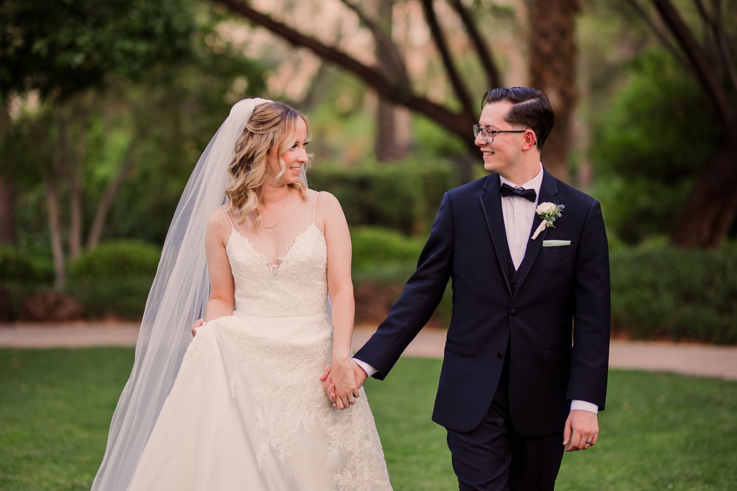 A couple in formal wedding attire walks together on a garden path surrounded by lush greenery at the JW Marriott Summerlin