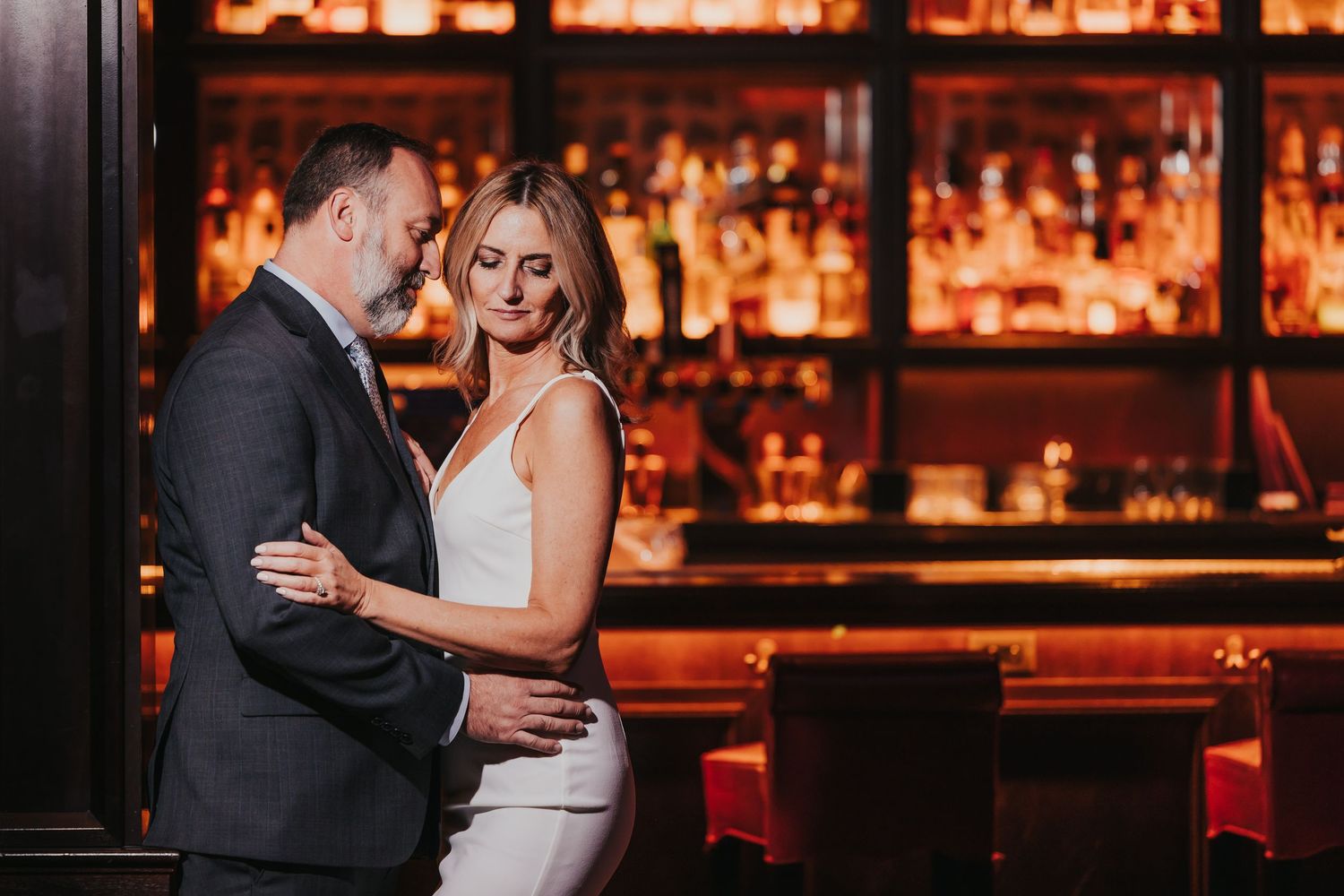 A couple shares a romantic dance in a dimly lit bar with warm amber lighting and red decor in the background.
