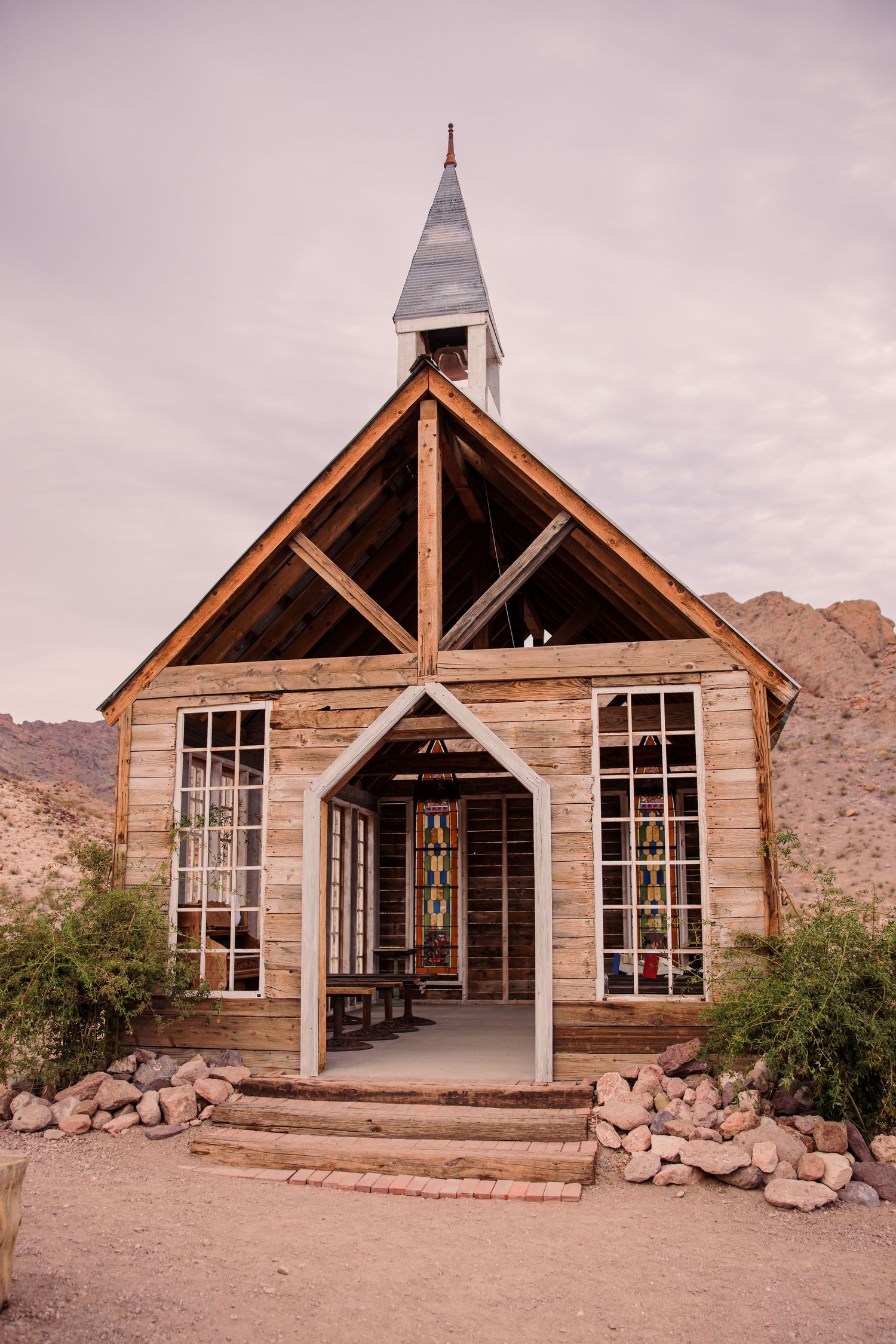 A rustic wooden chapel with large windows stands in a desert landscape against mountain backdrop.