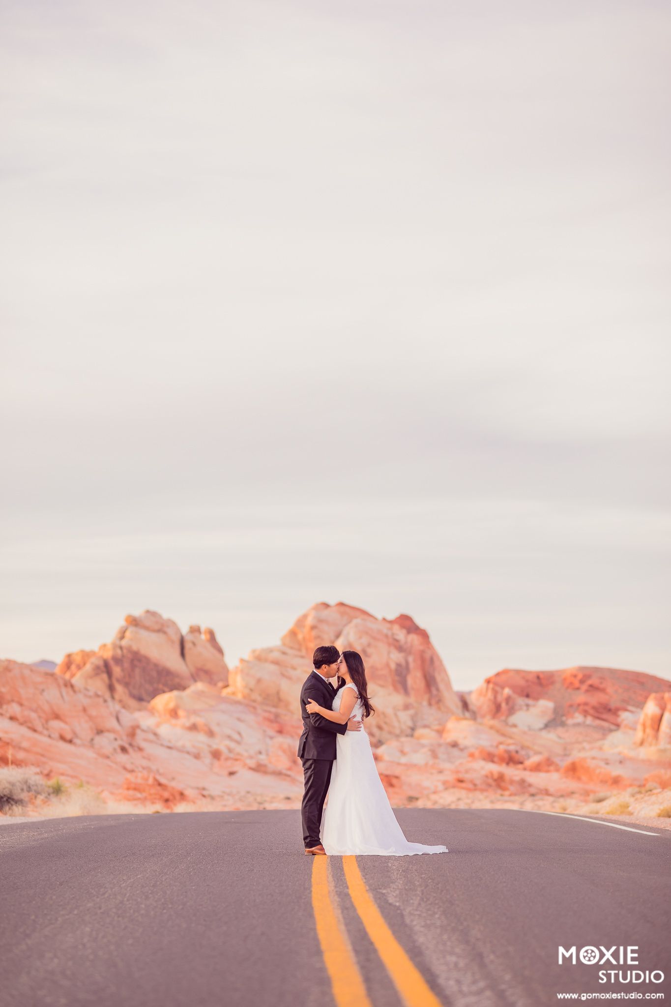 A couple embraces on an empty desert road with dramatic red rock formations and mountains in the background at sunset.