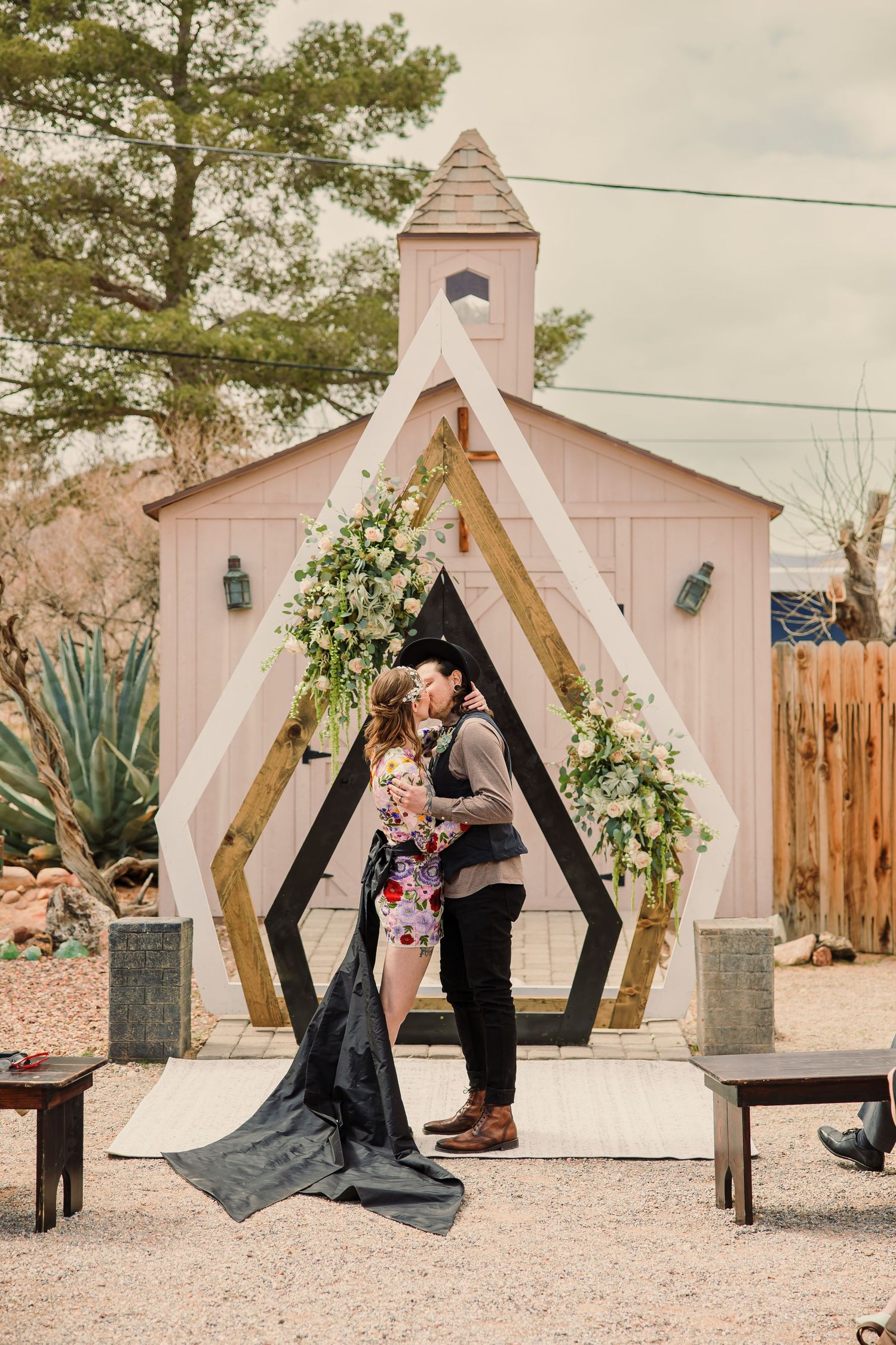 Modern geometric wooden wedding arch decorated with flowers against a pink chapel backdrop.