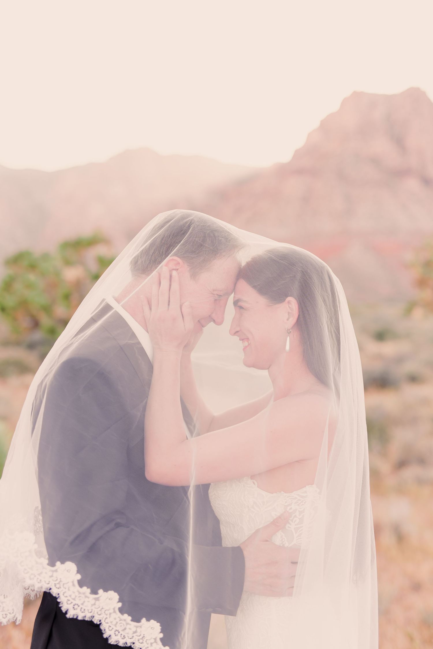 A romantic desert wedding moment as newlyweds share an intimate embrace under a flowing veil with mountains in background.