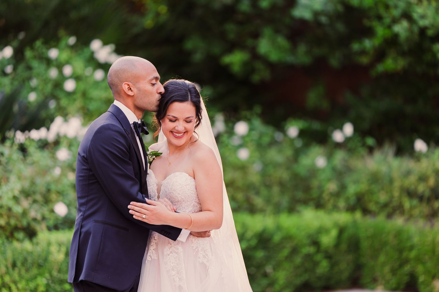 A couple in wedding attire share a romantic moment in a lush garden setting during their outdoor photo session.