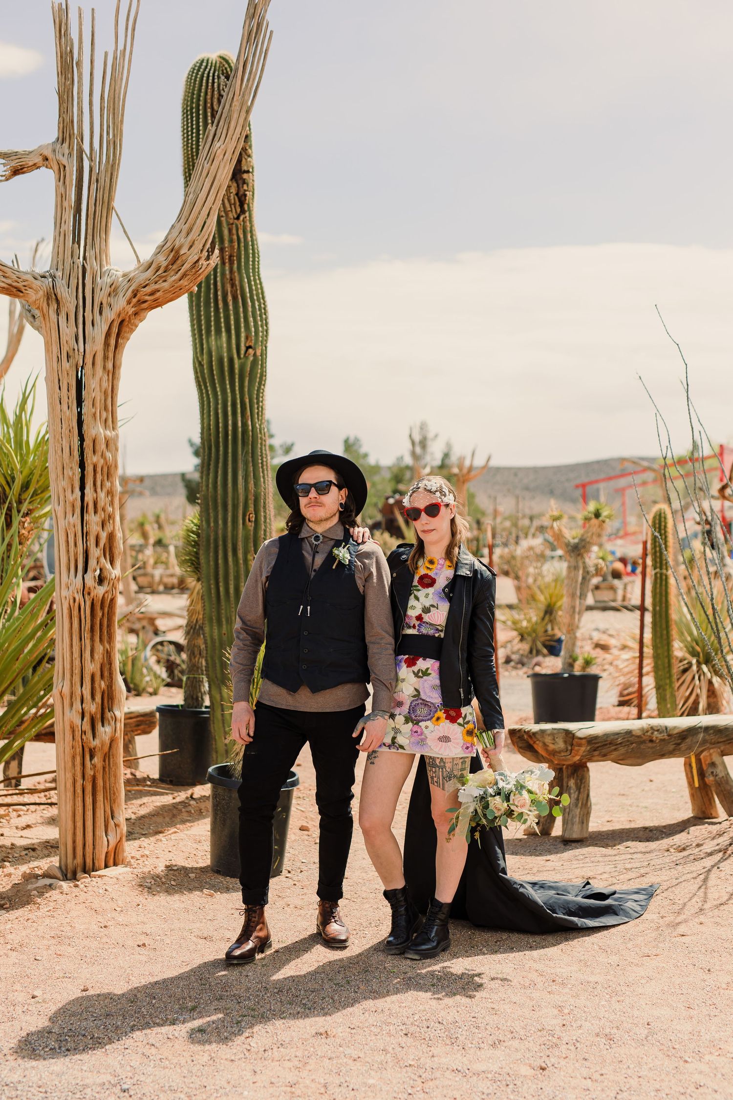 Two people pose together in a desert landscape with tall saguaro cacti and wooden posts in the background.