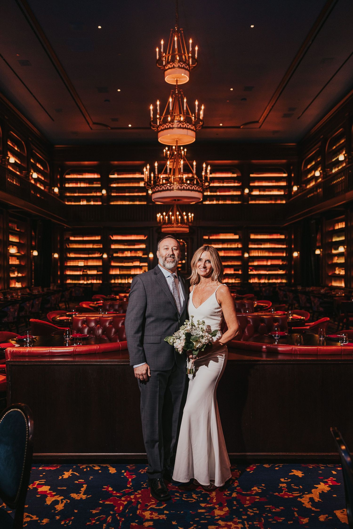 Couple poses in elegant dark bar with backlit bottles and chandeliers creating romantic ambiance for wedding photos.